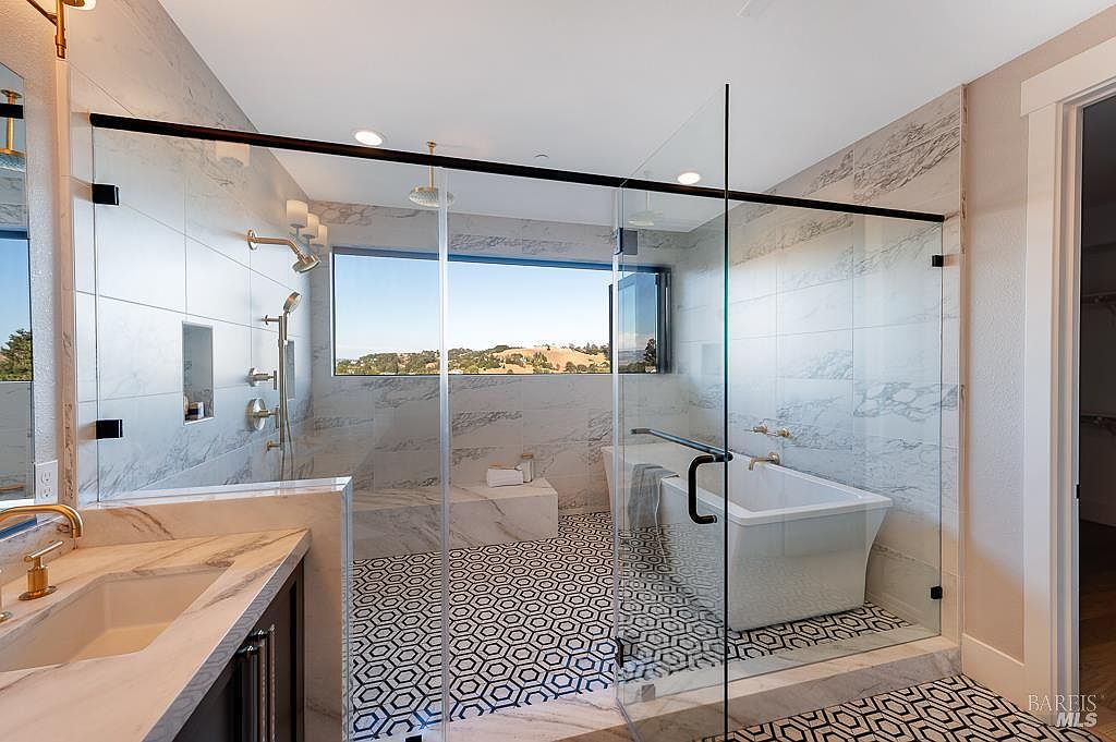 This luxurious primary bathroom features a seamless glass-enclosed wet room containing both a freestanding soaking tub and a spacious walk-in shower with marble-tiled walls. The room is anchored by a striking geometric patterned floor and a large window that provides a panoramic view of the surrounding hills. A sleek marble vanity with gold-toned fixtures completes the sophisticated, modern aesthetic.