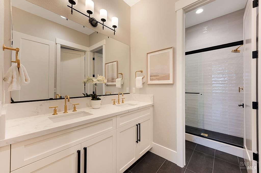 This elegant primary bathroom features a double vanity with white cabinetry, gold-toned fixtures, and a clean quartz countertop. A large, frameless mirror reflects the space, while a walk-in shower with sleek, horizontal subway tiles and a black base is visible through the doorway. The overall aesthetic is modern and sophisticated, characterized by a high-contrast color palette and refined finishes.