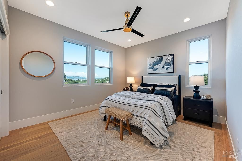 This bright and airy bedroom features a neutral color palette with light gray walls and warm hardwood flooring. The room is centered around a queen-sized bed with a striped duvet, flanked by two dark nightstands with matching lamps, and includes a small wooden bench at the foot of the bed. Large windows provide ample natural light and a scenic view, while a circular mirror and framed artwork add a touch of modern decor to the space.