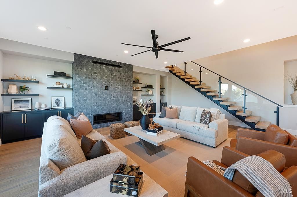 This spacious, modern living room features a striking floor-to-ceiling stone fireplace as its focal point, complemented by a contemporary open-tread staircase with glass railings. The room is furnished with a plush light-colored sofa, leather armchairs, and a minimalist coffee table, creating a warm and inviting atmosphere. The open-concept layout, neutral color palette, and high ceilings contribute to an airy, sophisticated aesthetic perfect for entertaining.
