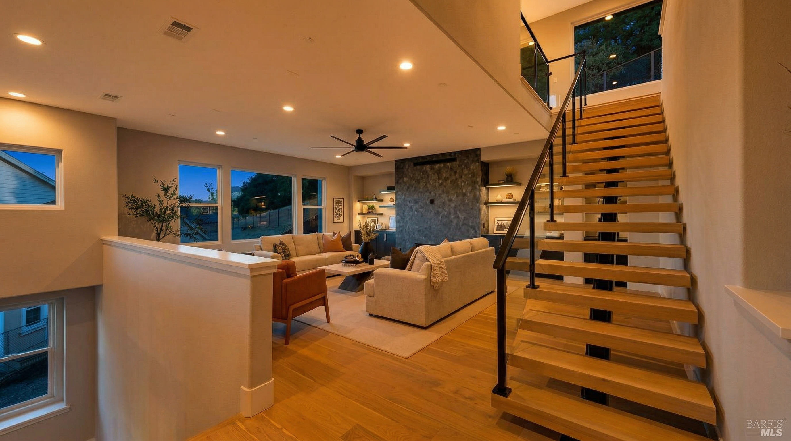 This modern open-concept living room features high ceilings, light hardwood floors, and a sophisticated floating staircase. A sleek, dark textured fireplace wall serves as the focal point, while the cozy seating area is oriented toward it, complemented by large windows that allow for plenty of natural light. The space combines contemporary design with warm, inviting textures for an elegant home atmosphere.