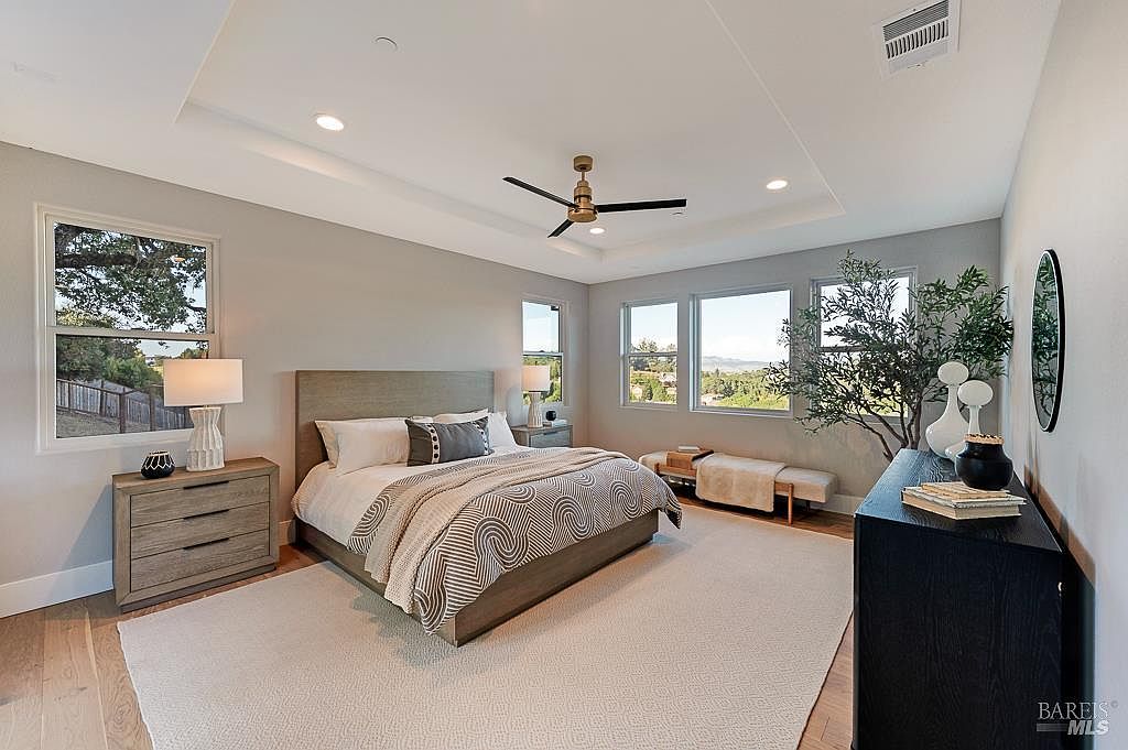 This spacious primary bedroom features a neutral color palette, a large bed with a textured headboard, and a patterned duvet cover. The room is illuminated by recessed lighting and a central ceiling fan, with large windows providing ample natural light and scenic views. A sleek black dresser and a wooden bench add functional elegance to the serene, well-appointed space.
