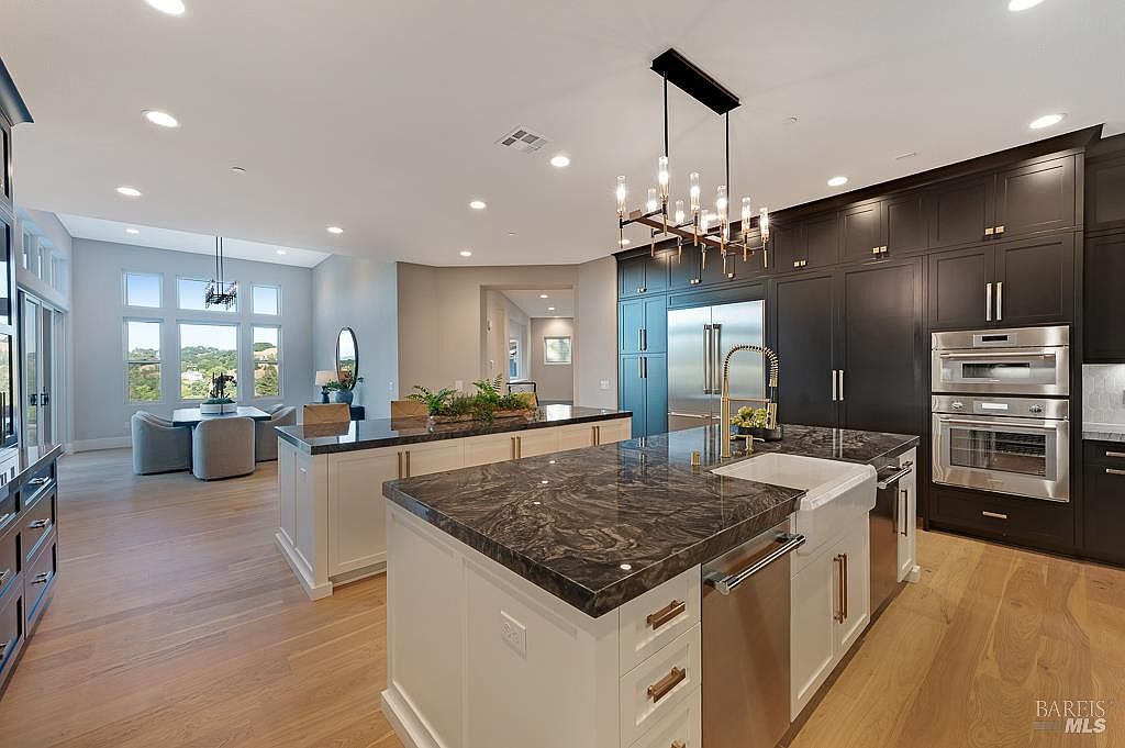 This expansive, high-end kitchen features a striking contrast between dark charcoal cabinetry and light-toned, marble-topped islands. The space is anchored by a large farmhouse sink, professional-grade stainless steel appliances, and a sophisticated linear chandelier hanging above the central workspace. With its open-concept layout flowing into a bright dining area and warm hardwood flooring, the room exudes a luxurious and inviting atmosphere.