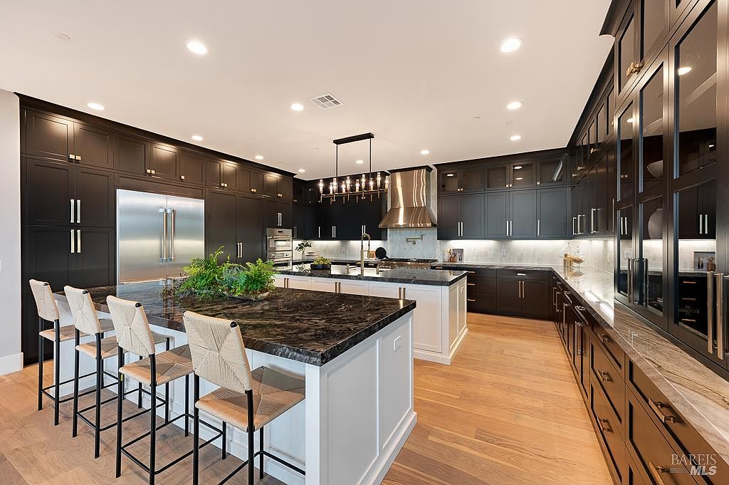 This sophisticated, high-end kitchen features a striking contrast between dark charcoal cabinetry and a bright white kitchen island, topped with dramatic, dark-veined stone countertops. The space is anchored by a large central island with woven bar stools, a professional-grade stainless steel range hood, and integrated appliances, all illuminated by a modern linear chandelier. The warm hardwood flooring and recessed lighting create an inviting, luxurious atmosphere perfect for both cooking and entertaining.