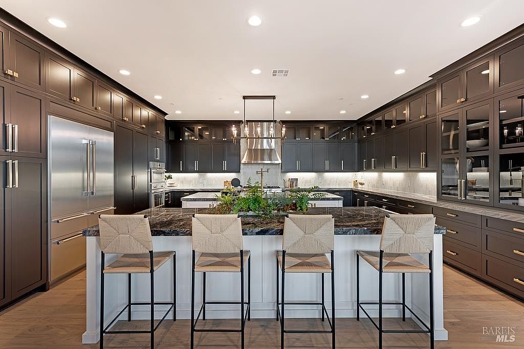 This sophisticated, high-end kitchen features dark cabinetry contrasted by a large, white-based central island topped with dramatic, dark veined stone. The space is anchored by a professional-grade stainless steel refrigerator and a sleek range hood, while four woven bar stools provide elegant seating. Recessed lighting and a stylish chandelier create a warm, inviting atmosphere, highlighting the seamless blend of modern luxury and functional design.