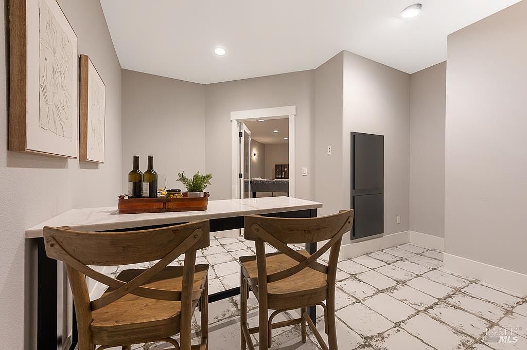 This cozy dining nook features a marble-topped table paired with two rustic wooden cross-back chairs, creating an inviting space for intimate meals. The room is accented by distressed white tile flooring and minimalist wall art, while a doorway leads into an adjacent room, adding depth to the perspective. The neutral color palette and clean lines provide a modern yet warm aesthetic suitable for a casual dining or breakfast area.