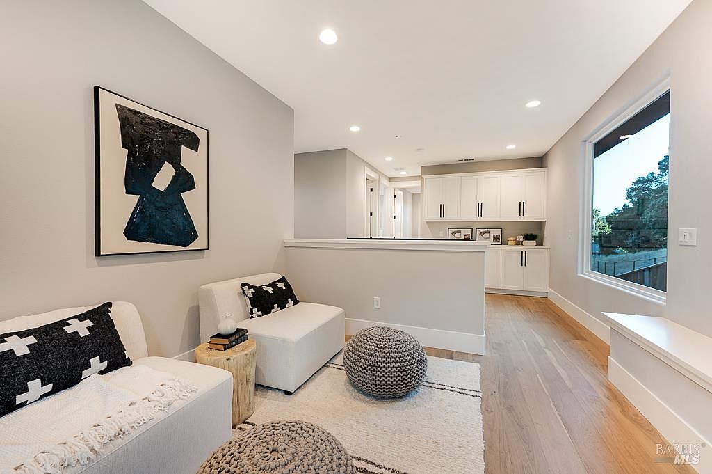 This bright and airy living room features a contemporary design with light hardwood floors, neutral walls, and a large window that invites natural light. The space is furnished with comfortable white seating, textured throw pillows, and a stylish gray pouf, all anchored by a plush area rug. A bold piece of abstract art serves as a focal point, while a half-wall separates the seating area from a clean, white-cabinetry kitchen space in the background.