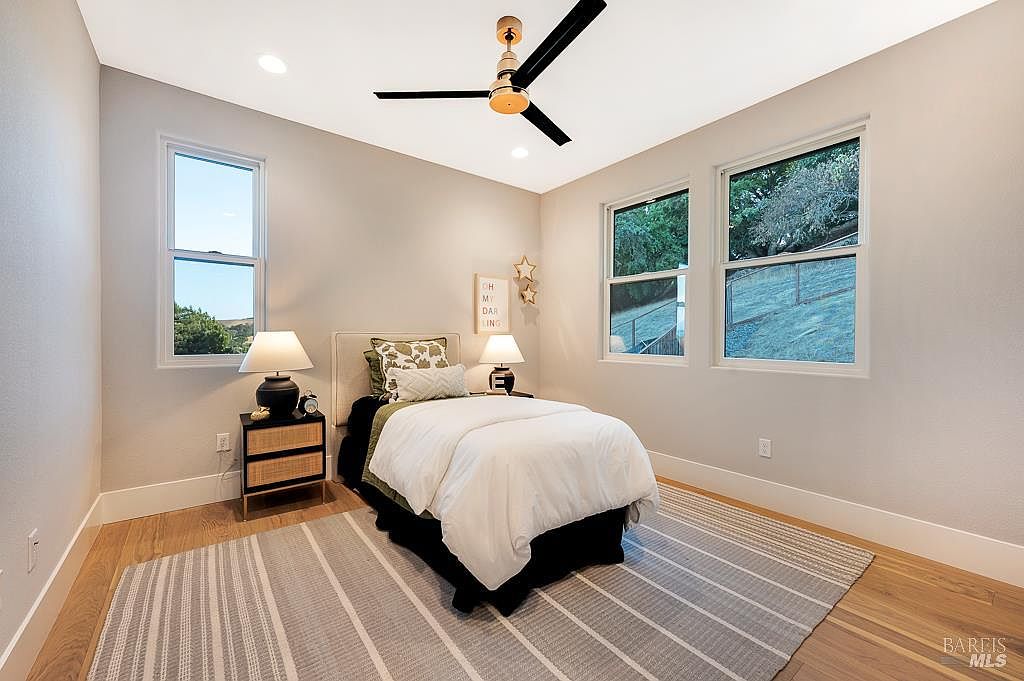 This bright and airy bedroom features a single bed centered against a neutral-toned wall, flanked by two matching nightstands with lamps. The room is accented by a modern black ceiling fan, a striped area rug, and large windows that provide ample natural light and a view of the outdoor landscape. The overall aesthetic is clean, contemporary, and inviting, making it an ideal space for guests or children.