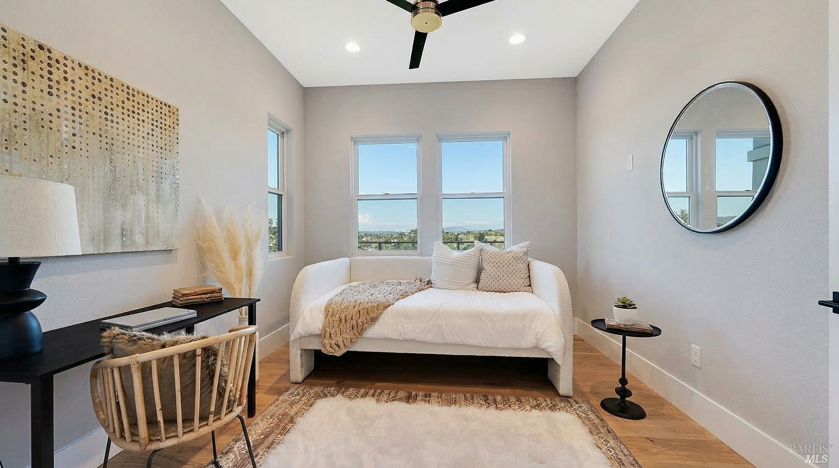 This versatile multipurpose room is styled as both a home office and a cozy secondary bedroom, featuring a daybed and a desk area. The room is bright and inviting with neutral walls, light wood flooring, and a large window overlooking a scenic view. It balances functional work space with comfortable seating, making it an ideal flexible-use room for a home.