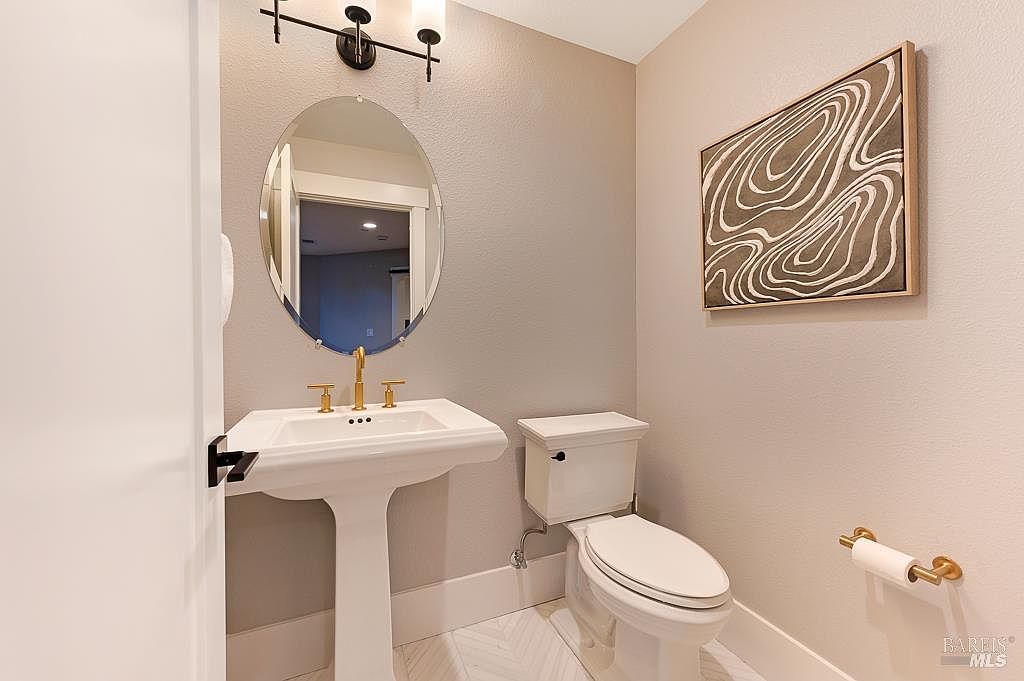 This elegant powder room features a classic pedestal sink with gold-toned fixtures, complemented by a sleek oval mirror and modern industrial-style lighting. The space is finished with neutral-toned walls, a white toilet, and a patterned tile floor, accented by a contemporary abstract art piece on the wall. The overall aesthetic is clean, sophisticated, and inviting, making it a perfect guest bathroom.