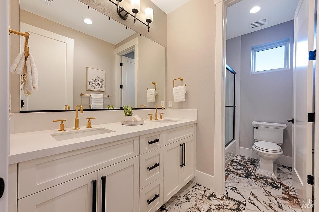 This modern primary bathroom features a double vanity with white cabinetry, gold-toned fixtures, and a sleek quartz countertop. The space is accented by a large mirror, marble-patterned flooring, and a separate water closet area with a shower. The overall aesthetic is bright, clean, and luxurious, offering a contemporary feel.