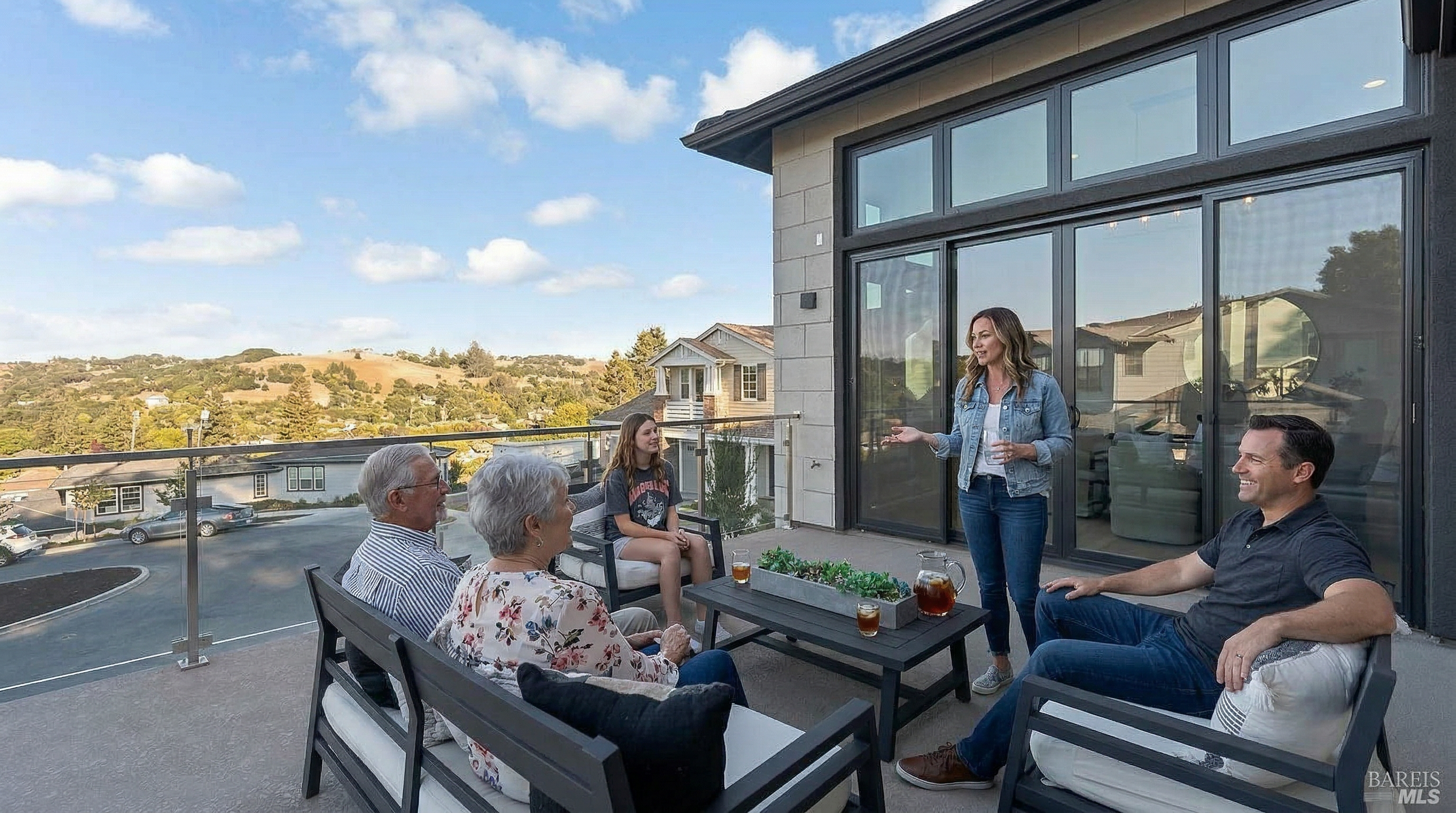 This modern, elevated balcony offers a spacious outdoor living area furnished with contemporary dark-framed seating and a central coffee table. The space features a sleek glass railing that provides an unobstructed, panoramic view of the surrounding neighborhood and rolling hills. Large sliding glass doors connect the balcony to the interior, creating a seamless transition between indoor and outdoor living spaces.