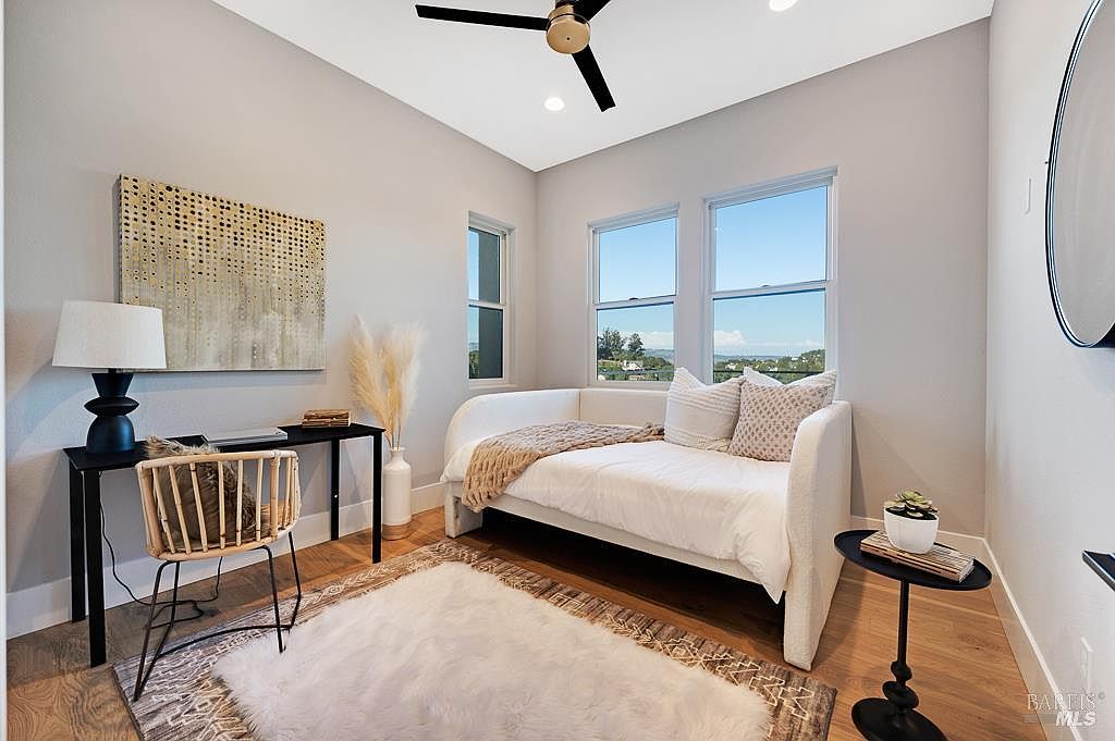 This versatile room functions as a bright and airy home office or guest bedroom, featuring a daybed with neutral bedding and a sleek black desk setup. The space is accented by a textured area rug, a modern ceiling fan, and large windows that provide ample natural light and a scenic view. The overall aesthetic is clean, contemporary, and inviting, making it a flexible space for work or relaxation.