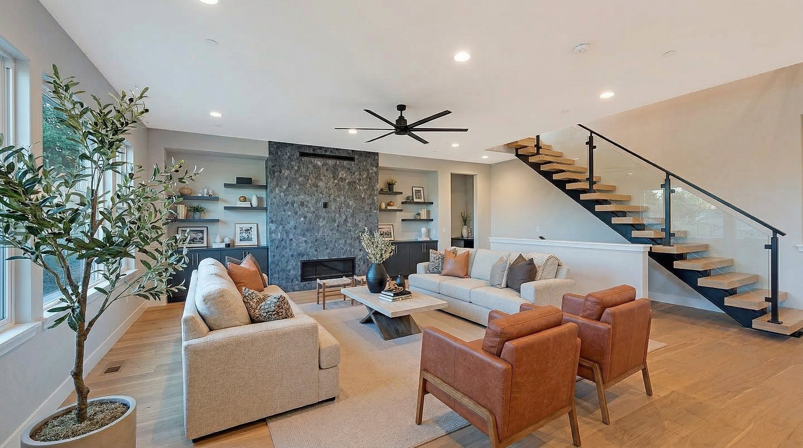This spacious, open-concept living room features a striking floor-to-ceiling textured stone fireplace focal point and a modern floating staircase with glass railings. The airy space is furnished with two beige sofas, two cognac leather accent chairs, and an olive tree, creating a warm and contemporary atmosphere. Large windows and recessed lighting keep the living area bright, making it an inviting space for relaxation or entertaining.