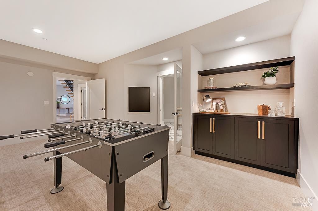 This finished basement space features a foosball table as the central focal point, set against a neutral color palette with plush carpeting. To the right, a built-in wet bar with dark cabinetry, gold hardware, and open shelving provides a sophisticated area for entertaining. The room is well-lit with recessed lighting and offers a clear view into an adjacent hallway and doorway, creating an inviting and functional recreational area.