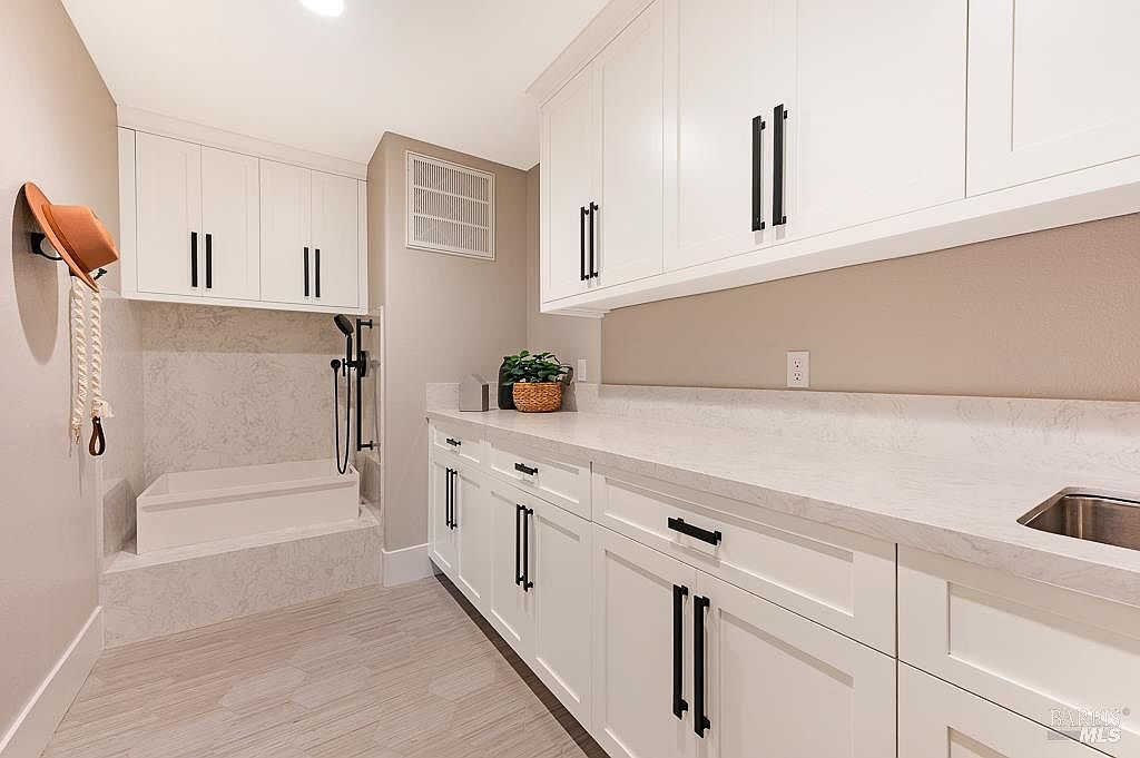 This bright and functional laundry room features crisp white cabinetry with modern black hardware and expansive quartz countertops. A built-in pet washing station with a handheld sprayer is tucked into the corner, complemented by light-toned, wood-look tile flooring. The space is clean and contemporary, offering a highly organized and versatile area for household chores.