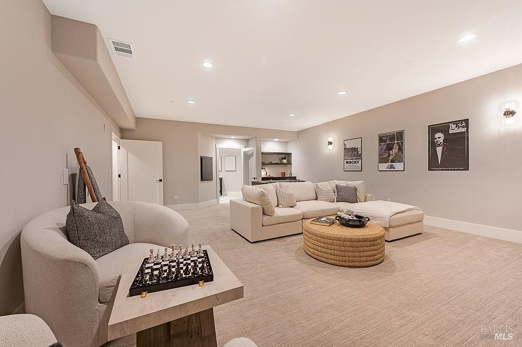 This spacious, modern basement features a comfortable lounge area with a large sectional sofa, a textured round ottoman, and a curved accent chair paired with a chess set. The neutral color palette, recessed lighting, and framed movie posters create a cozy, cinematic atmosphere perfect for entertainment. The open layout flows seamlessly toward a small wet bar area in the background, highlighting the home's versatile living space.