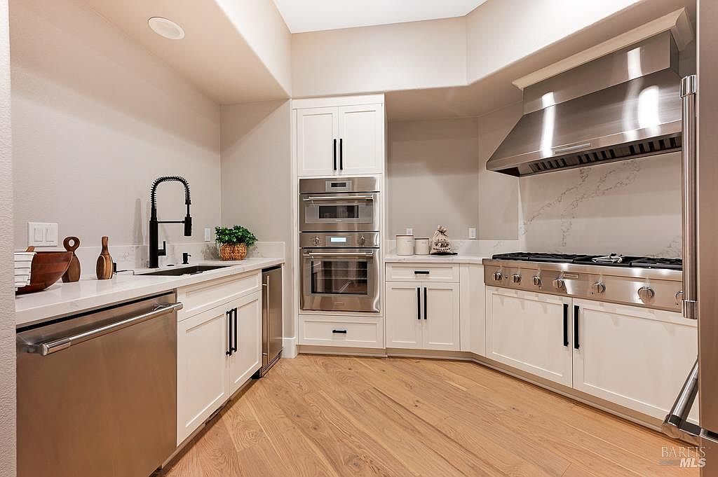 This modern, high-end kitchen features crisp white cabinetry, sleek stainless steel appliances, and elegant quartz countertops. The space is anchored by a professional-grade range with a large stainless steel hood, complemented by warm hardwood flooring and a contemporary black faucet. The perspective is a wide-angle shot that highlights the functional layout and clean, sophisticated aesthetic of the culinary space.