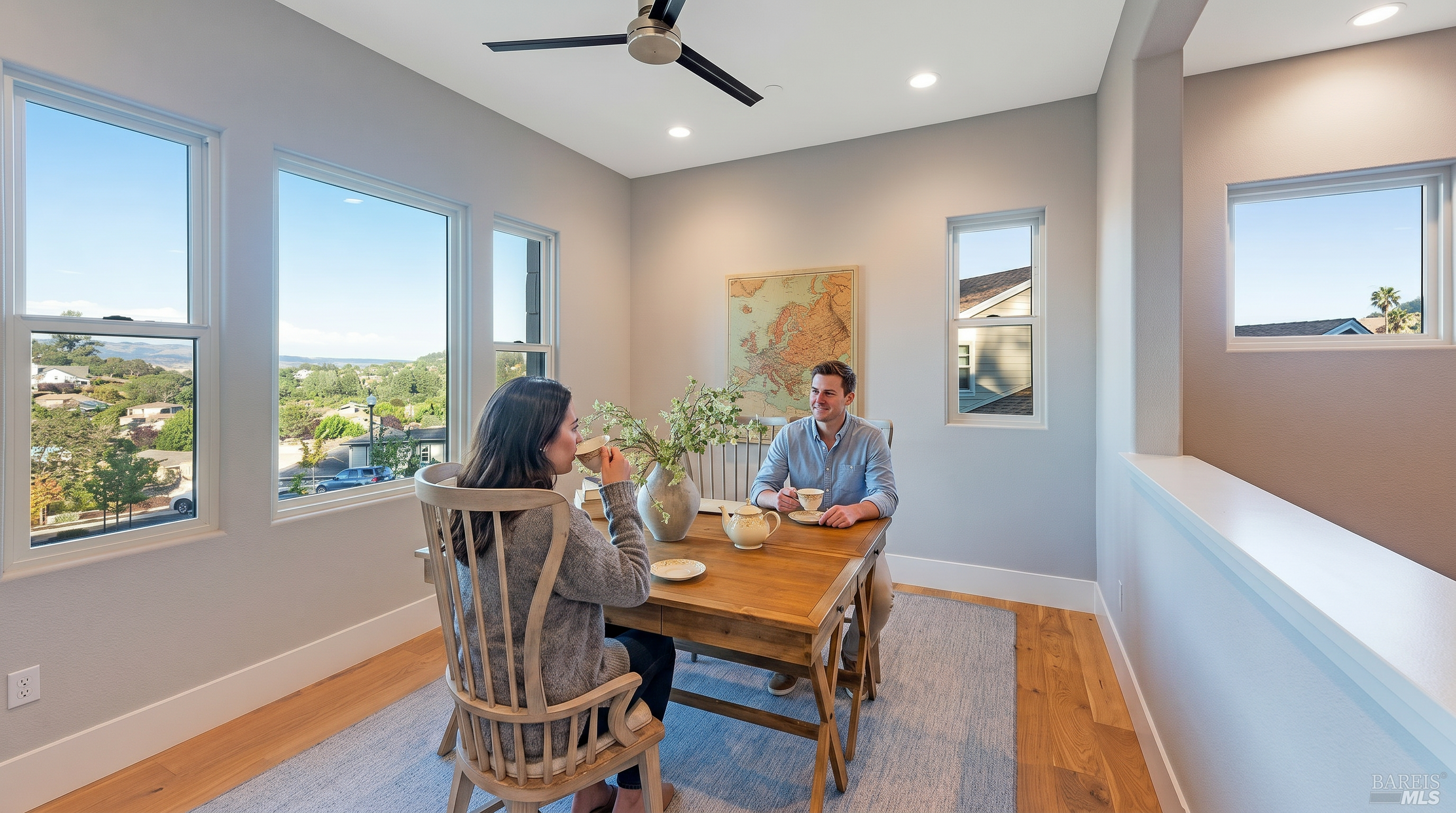 This bright and airy home office features a rustic wooden desk centered on a textured area rug, complemented by a classic wooden chair and a decorative vase of greenery. Large windows provide an expansive view of the surrounding neighborhood, while a vintage-style map on the wall adds a touch of character to the neutral-toned space. The room is illuminated by recessed lighting and a modern ceiling fan, creating a clean, productive, and inviting atmosphere.