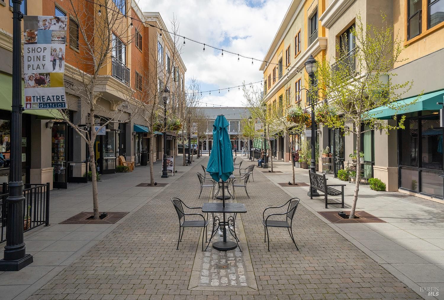 This image captures a charming, pedestrian-friendly outdoor shopping and dining promenade, characterized by its brick-paved walkway and symmetrical architecture. The scene features inviting bistro-style seating, mature trees, and string lights overhead, creating a vibrant and welcoming atmosphere for residents and visitors. The perspective is a centered, eye-level shot looking down the corridor, emphasizing the clean, well-maintained urban design of the district.