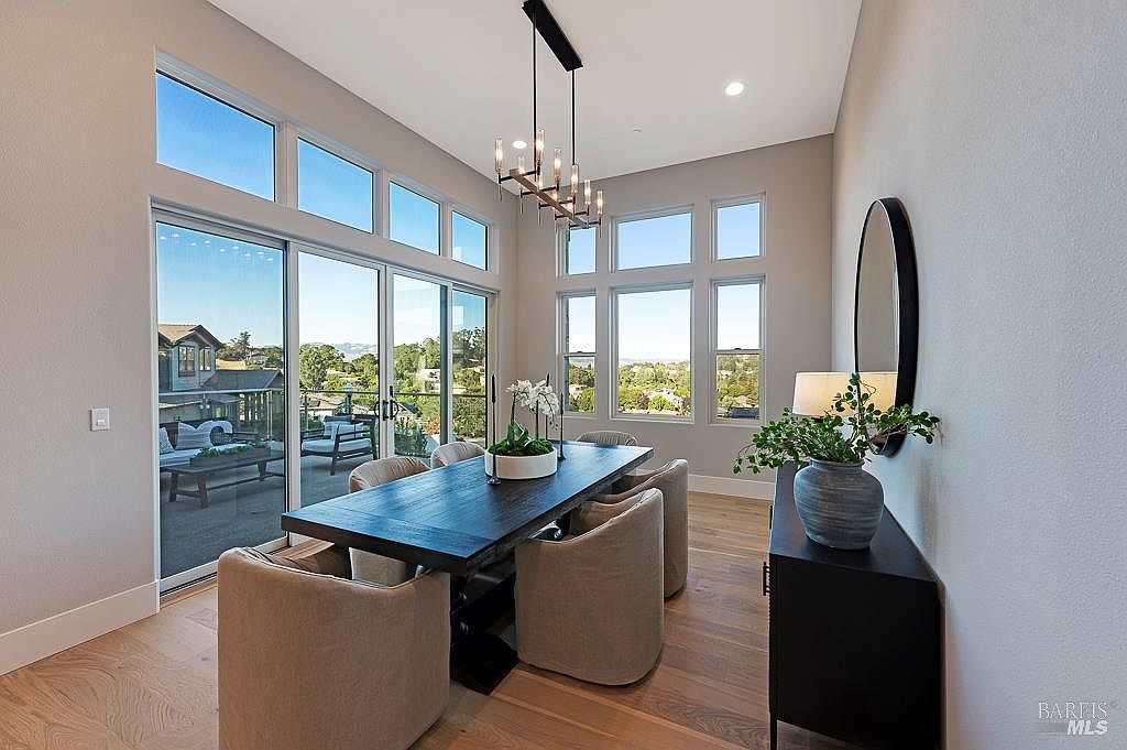 This elegant dining room features a dark wood table surrounded by plush, light-colored upholstered chairs, creating a sophisticated contrast. A modern, multi-light chandelier hangs above the table, while large windows and sliding glass doors provide abundant natural light and scenic views of the surrounding landscape. The space is accented by a sleek black sideboard, a circular mirror, and a decorative plant, offering a refined and inviting atmosphere.