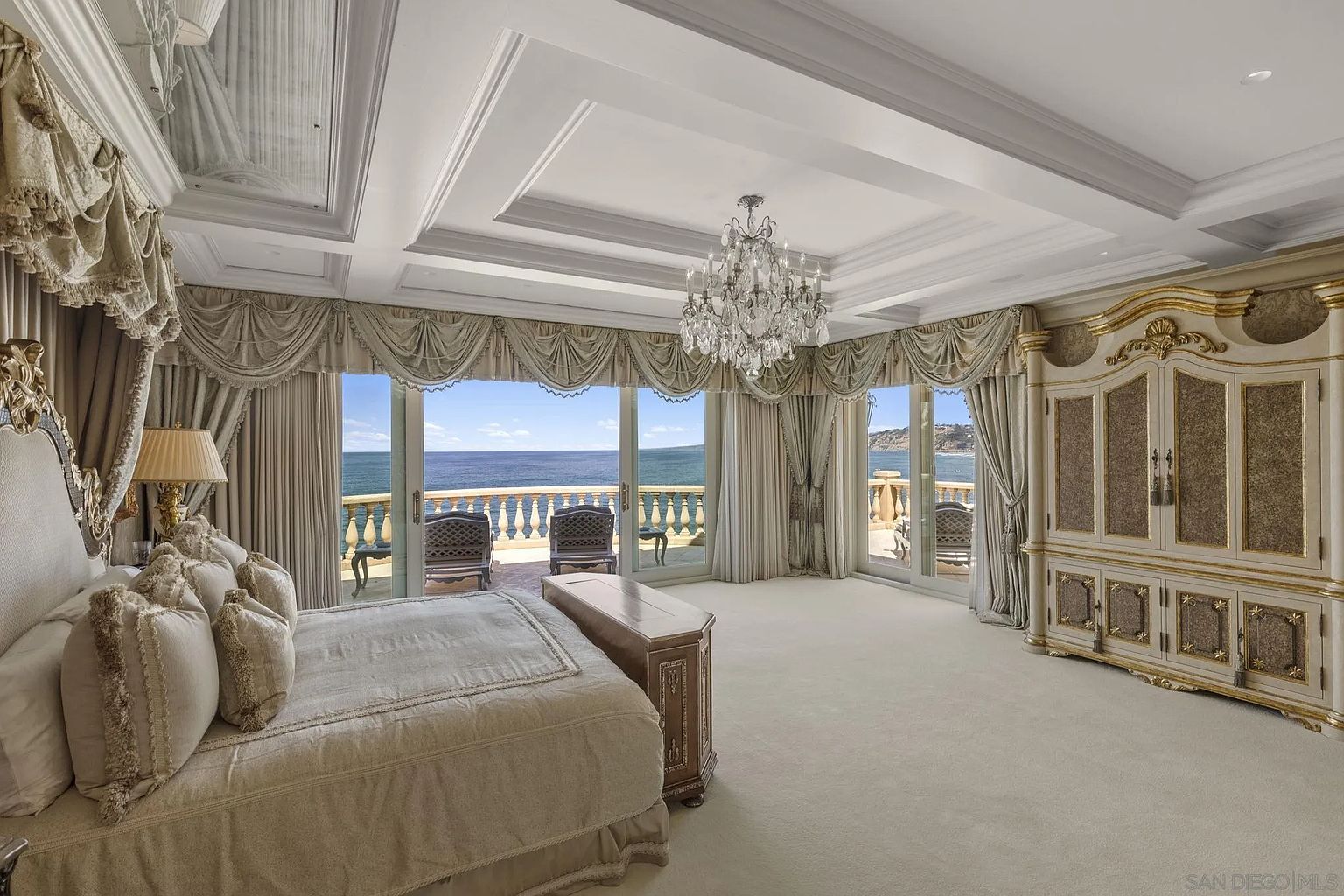 This is a luxurious primary bedroom featuring a large bed with ornate detailing and plush bedding. The room boasts floor-to-ceiling windows offering stunning ocean views, complemented by elegant drapery. A crystal chandelier hangs from the coffered ceiling, and a large, decorative armoire stands against one wall, adding to the room's opulent and spacious feel.