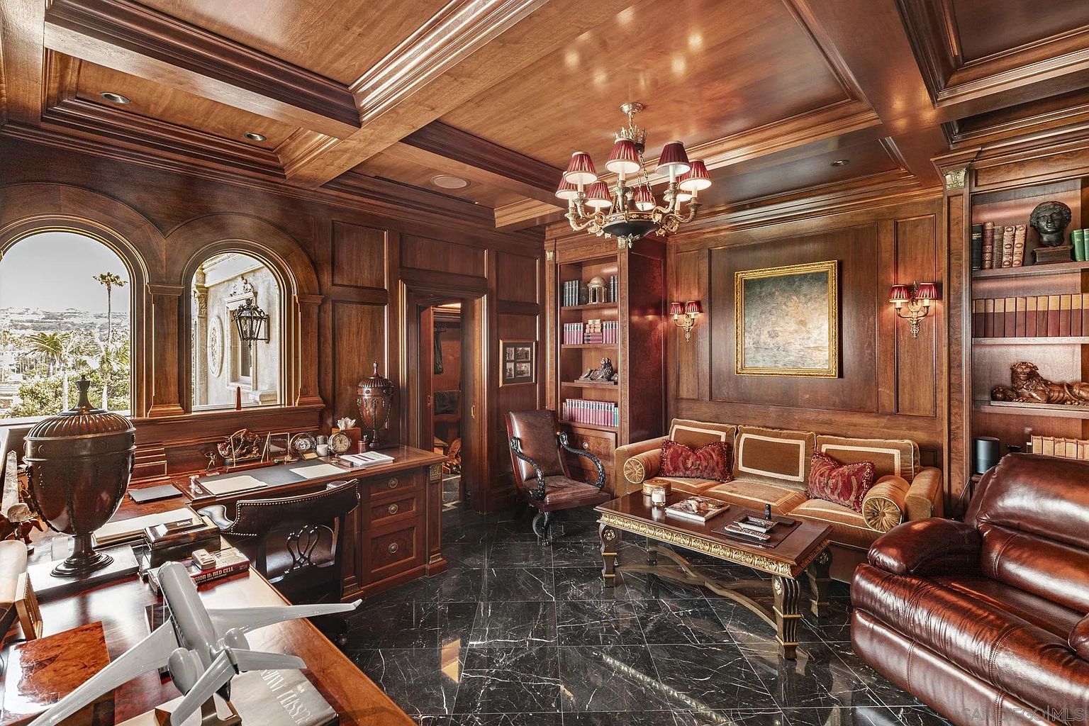 This is a luxurious and stately home office featuring rich wood paneling, a black marble floor, and a coffered wood ceiling. The room is furnished with a large wooden desk, leather chairs, a sofa, and built-in bookshelves. A chandelier provides warm lighting, and a large window offers a view to the outside. The perspective is from the front of the desk, looking into the room.
