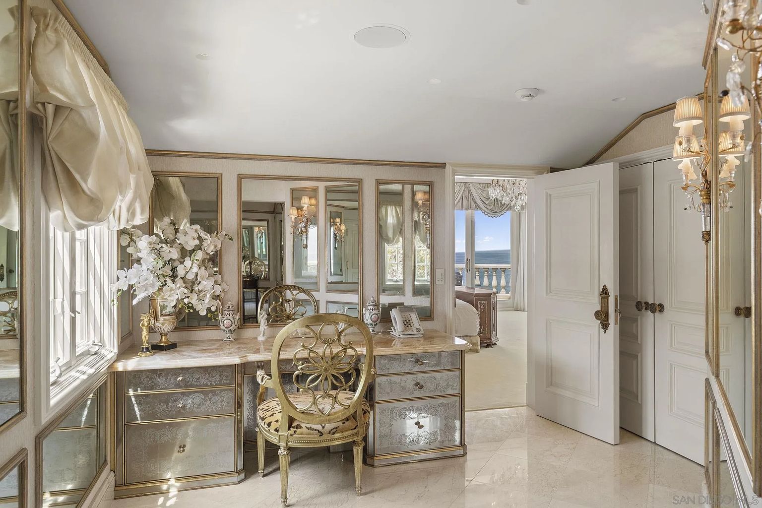 This is a luxurious primary bedroom featuring a vanity area with multiple mirrors and an ornate chair. The room has a sophisticated design with elegant furnishings, including a chandelier visible through an open doorway leading to a balcony with ocean views. The color palette is soft and neutral, creating a serene and upscale atmosphere.