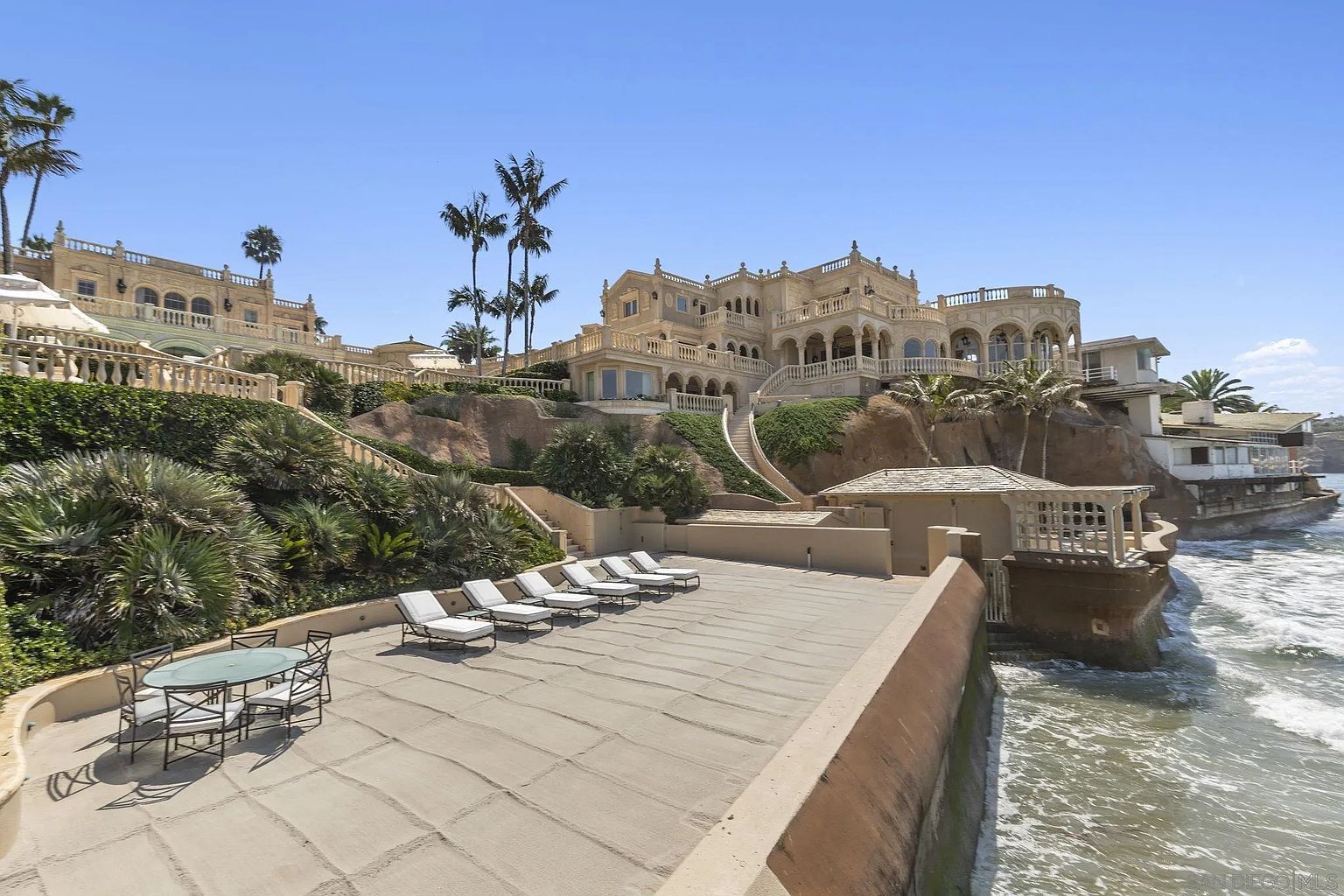 This exterior shot showcases a luxurious patio area with lounge chairs and a dining set, set against the backdrop of a grand mansion. The patio overlooks the ocean, offering stunning views and a sense of exclusivity. The architectural style of the mansion is reminiscent of Mediterranean design, with multiple levels, balconies, and intricate detailing.