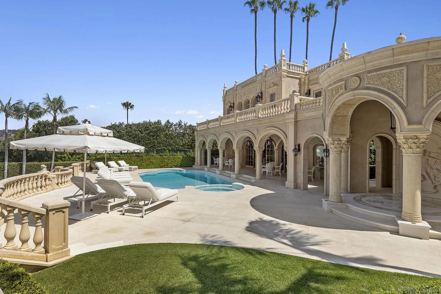 This image showcases a luxurious outdoor pool and spa area adjacent to a grand estate. The pool is surrounded by elegant lounge chairs and a large umbrella, while the architecture features arched walkways, ornate columns, and a second-story balcony. Lush green grass and palm trees add to the opulent and resort-like atmosphere.