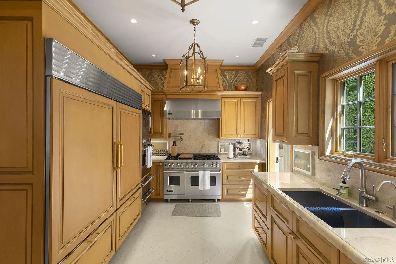 This is a well-lit kitchen featuring custom wood cabinetry, stainless steel appliances, and a light-colored tile floor. The kitchen includes a large refrigerator, a professional-grade range, and a sink with a window view. The overall style is traditional and luxurious, with a focus on high-end finishes and functionality.