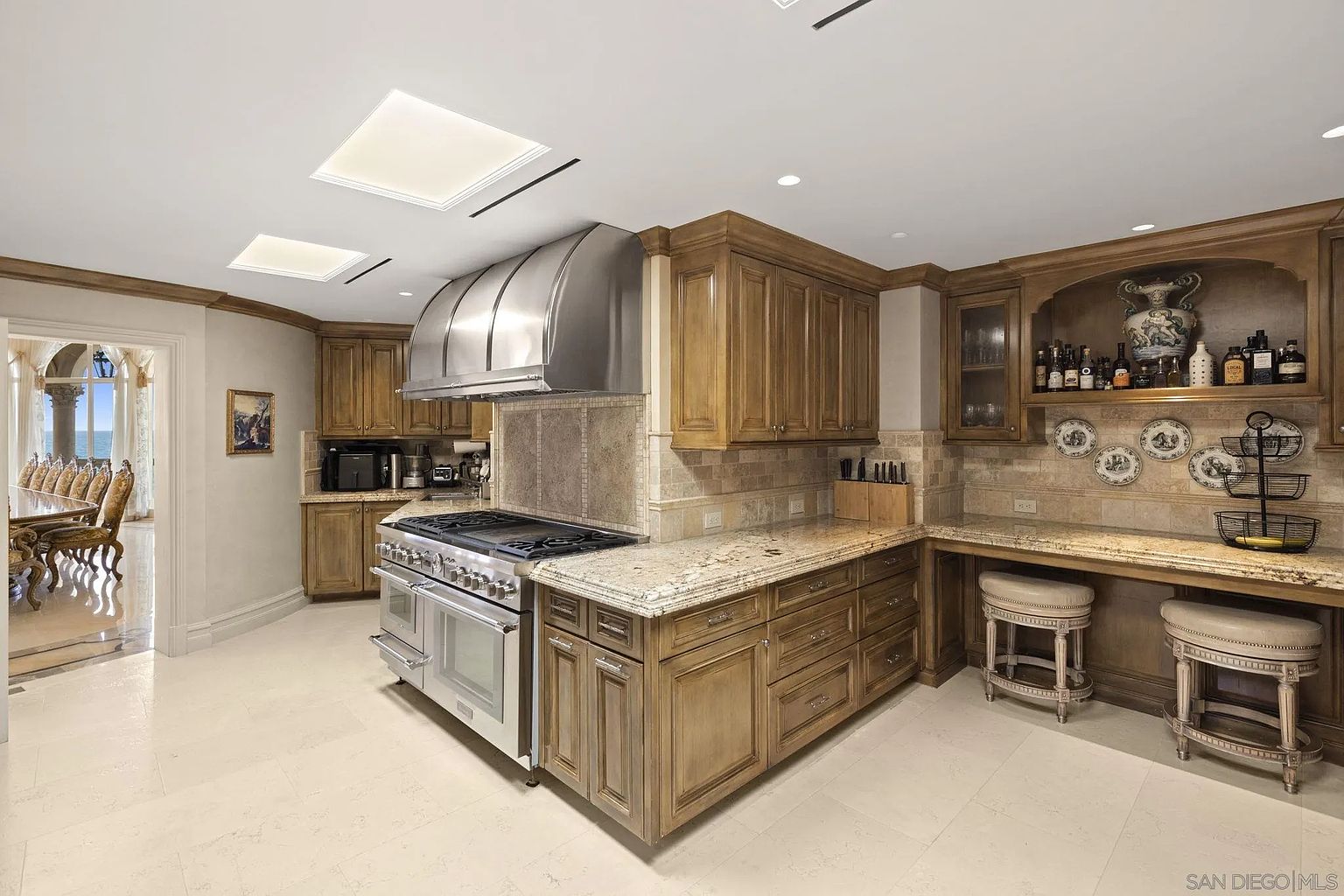 This is a well-appointed kitchen featuring custom wood cabinetry, granite countertops, and stainless steel appliances, including a professional-grade range with a large vent hood. The kitchen also includes a built-in bar area with seating, decorative plates, and ample storage. Natural light streams in from skylights, enhancing the warm and inviting atmosphere.