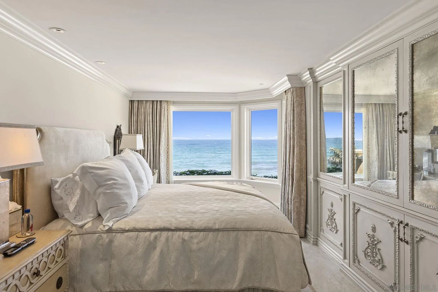 This is a luxurious primary bedroom featuring a large bed with plush white bedding, a bay window offering ocean views, and custom built-in cabinetry with mirrored doors. The room is decorated in a neutral color palette with elegant architectural details, creating a serene and upscale atmosphere. The room is shot at eye level to show the size of the room.