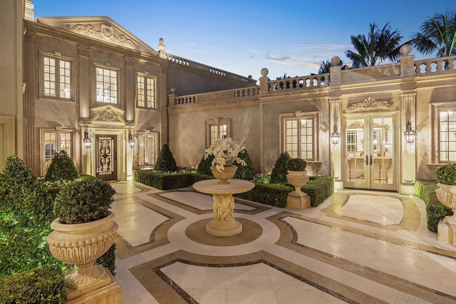 This is a luxurious entryway to a grand estate, featuring a meticulously designed courtyard with a central fountain and ornate landscaping. The architecture showcases classical elements with detailed stonework, symmetrical window placement, and elegant lighting fixtures. The overall impression is one of opulence and sophisticated design, creating a welcoming and impressive approach to the property.