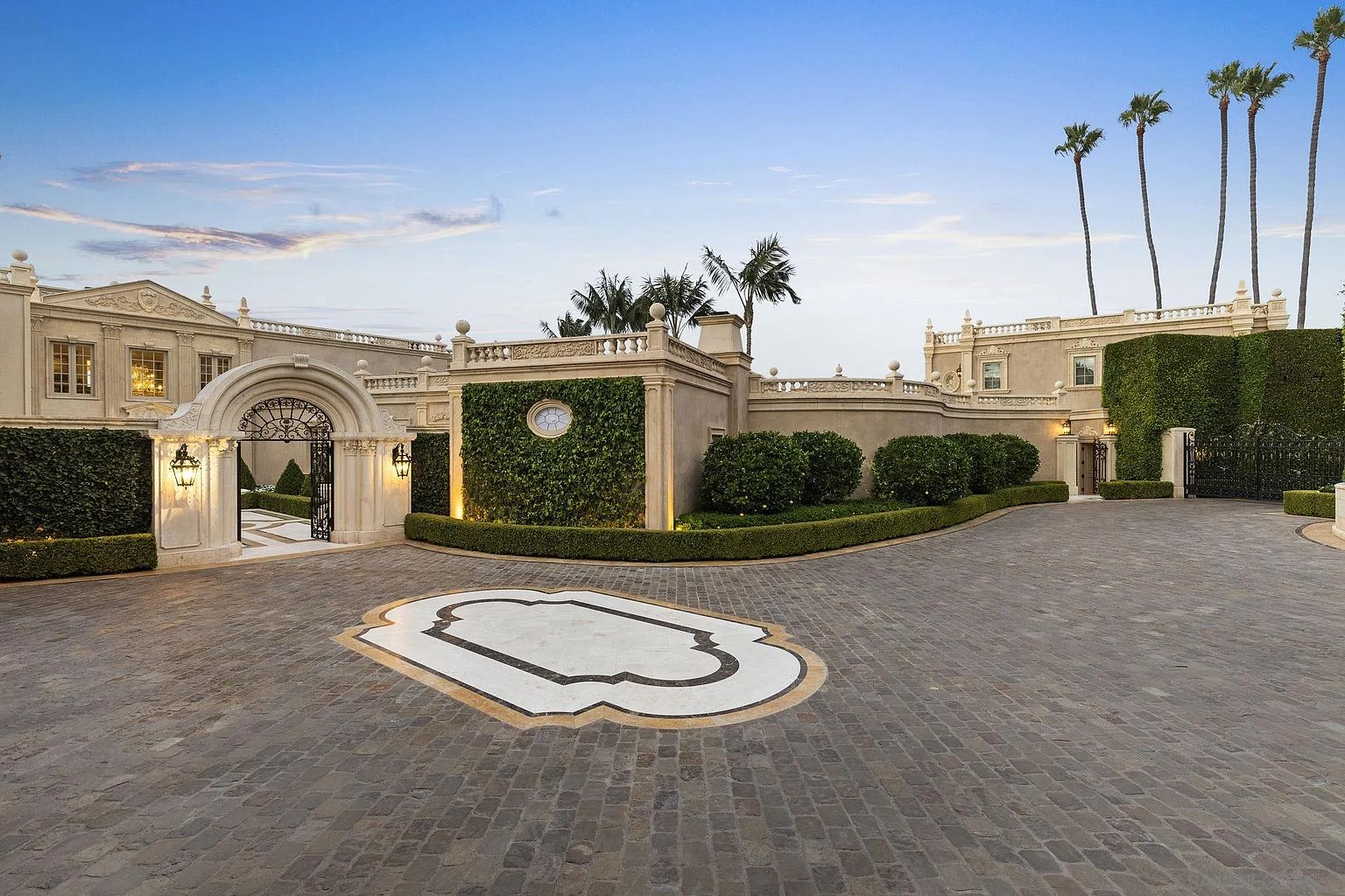 This is a grand front view of a luxurious estate, showcasing a circular driveway paved with cobblestone and a decorative central design. The property features manicured hedges, ornate gates, and architectural details such as arched entryways and decorative balustrades. Tall palm trees punctuate the skyline, adding to the property's upscale and elegant ambiance.