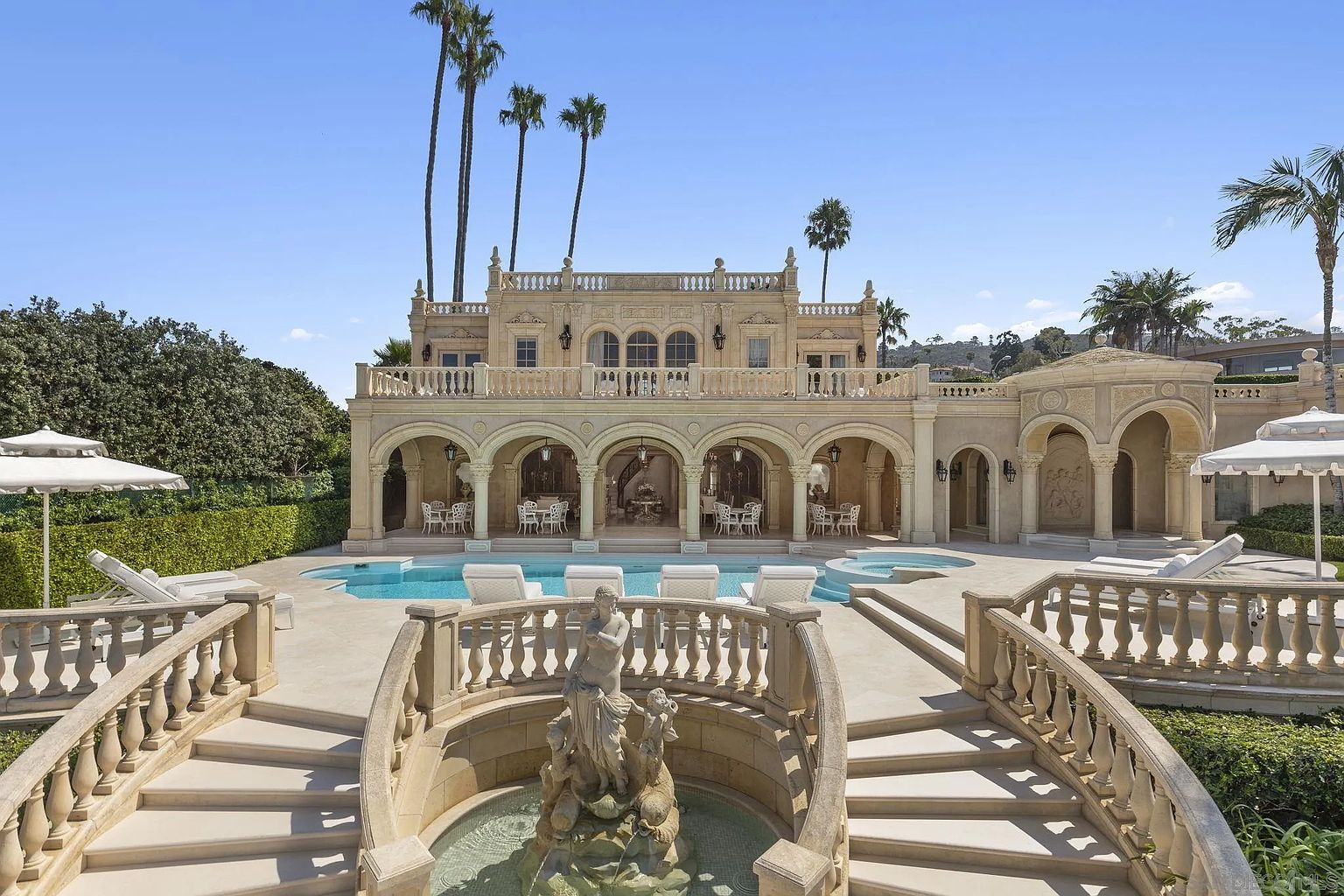 This image showcases the rear exterior of a luxurious estate, highlighting a stunning pool and spa area. The architecture features elegant arches, a second-story balcony, and a grand fountain with a statue, all contributing to a sophisticated and opulent ambiance. Palm trees dot the landscape, enhancing the property's appeal and creating a resort-like atmosphere.