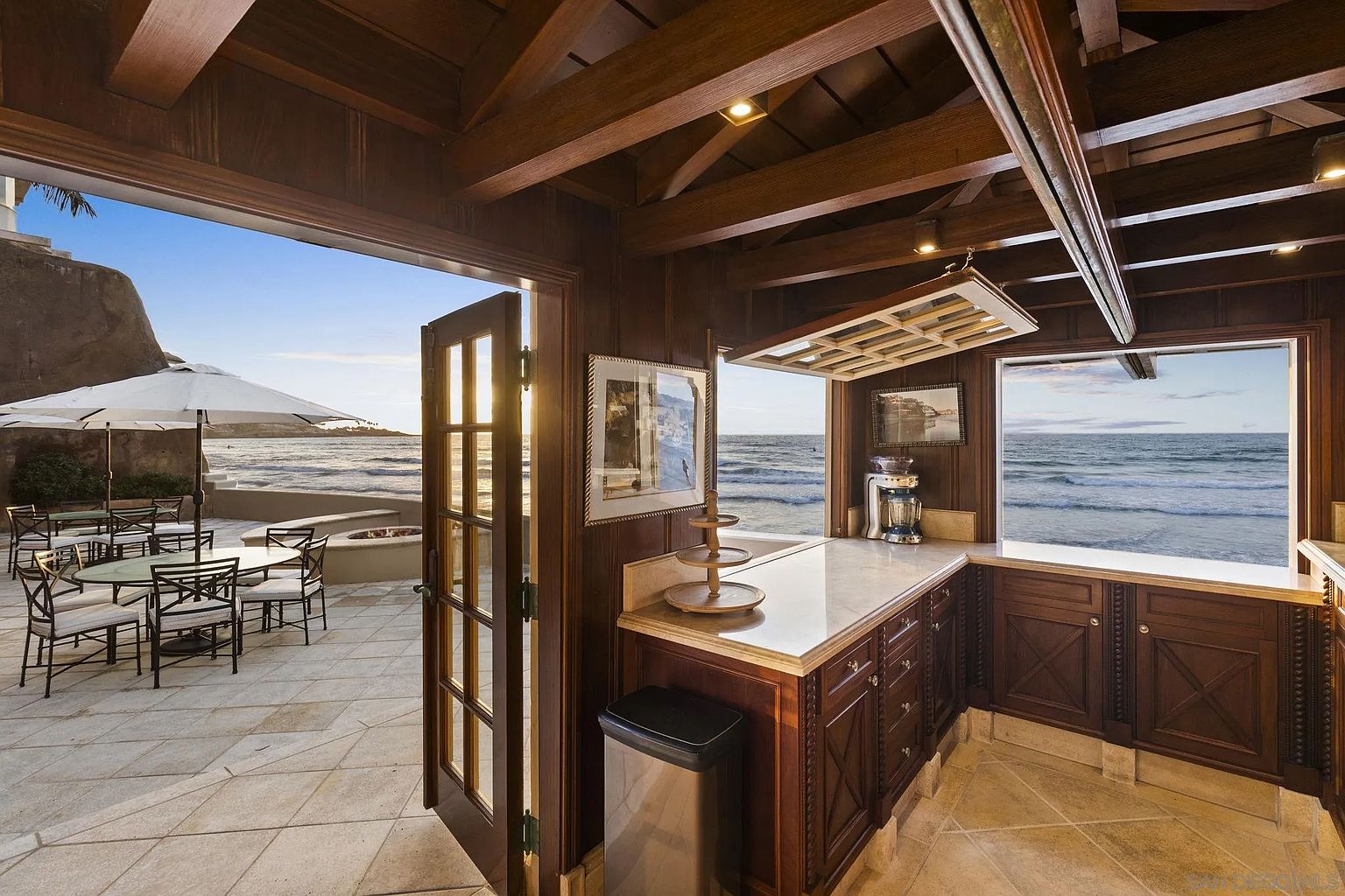 This image showcases a unique indoor/outdoor kitchen space with rich wood cabinetry and a marble countertop. The kitchen opens onto a patio overlooking the ocean, creating a seamless transition between indoor and outdoor living. The design emphasizes natural light and stunning views, making it an ideal space for entertaining or relaxing.