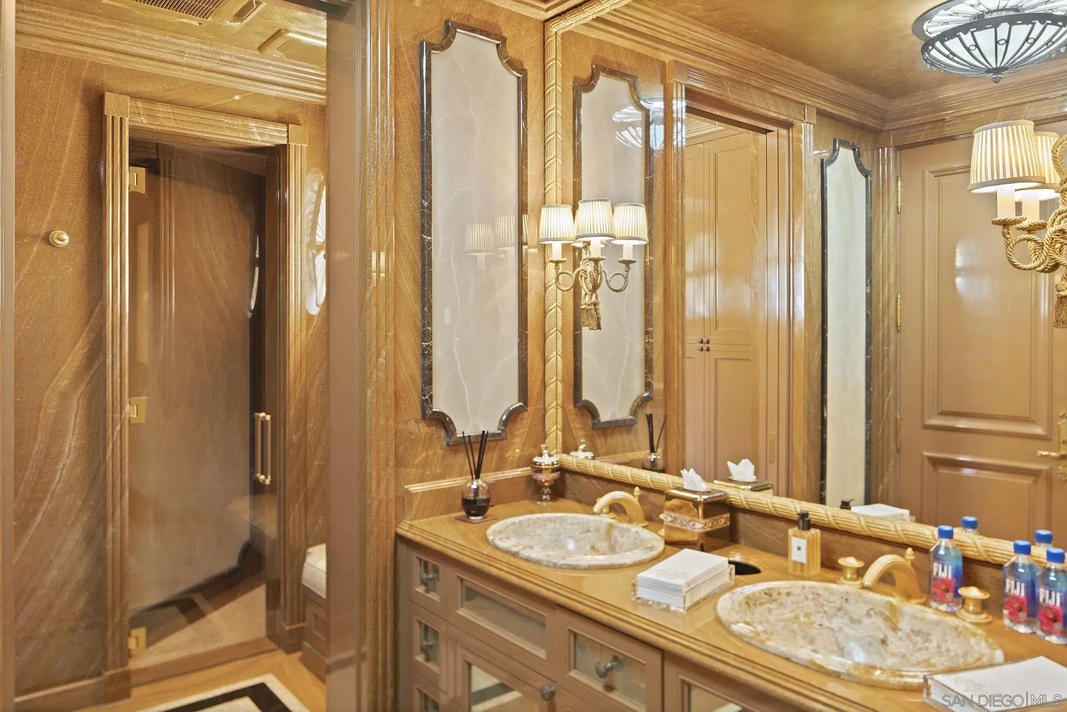 This is a luxurious primary bathroom featuring dual sinks with marble countertops and ornate gold fixtures. The walls are covered in a textured gold material, complemented by decorative mirrors and sconces. A glass-enclosed shower is visible to the left, adding to the opulent and well-appointed feel of the space.