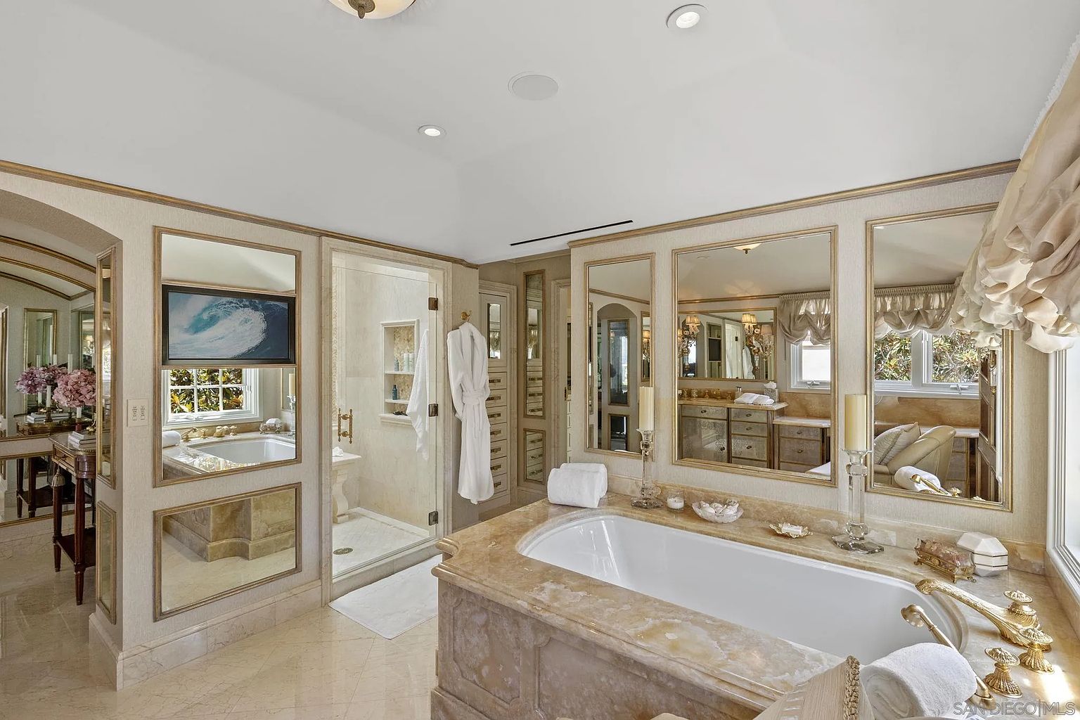 This is a luxurious primary bathroom featuring a marble-encased bathtub as the focal point. The room is adorned with multiple mirrors, enhancing the sense of space and light. A glass-enclosed shower is visible to the left, and the overall design exudes elegance and sophistication.
