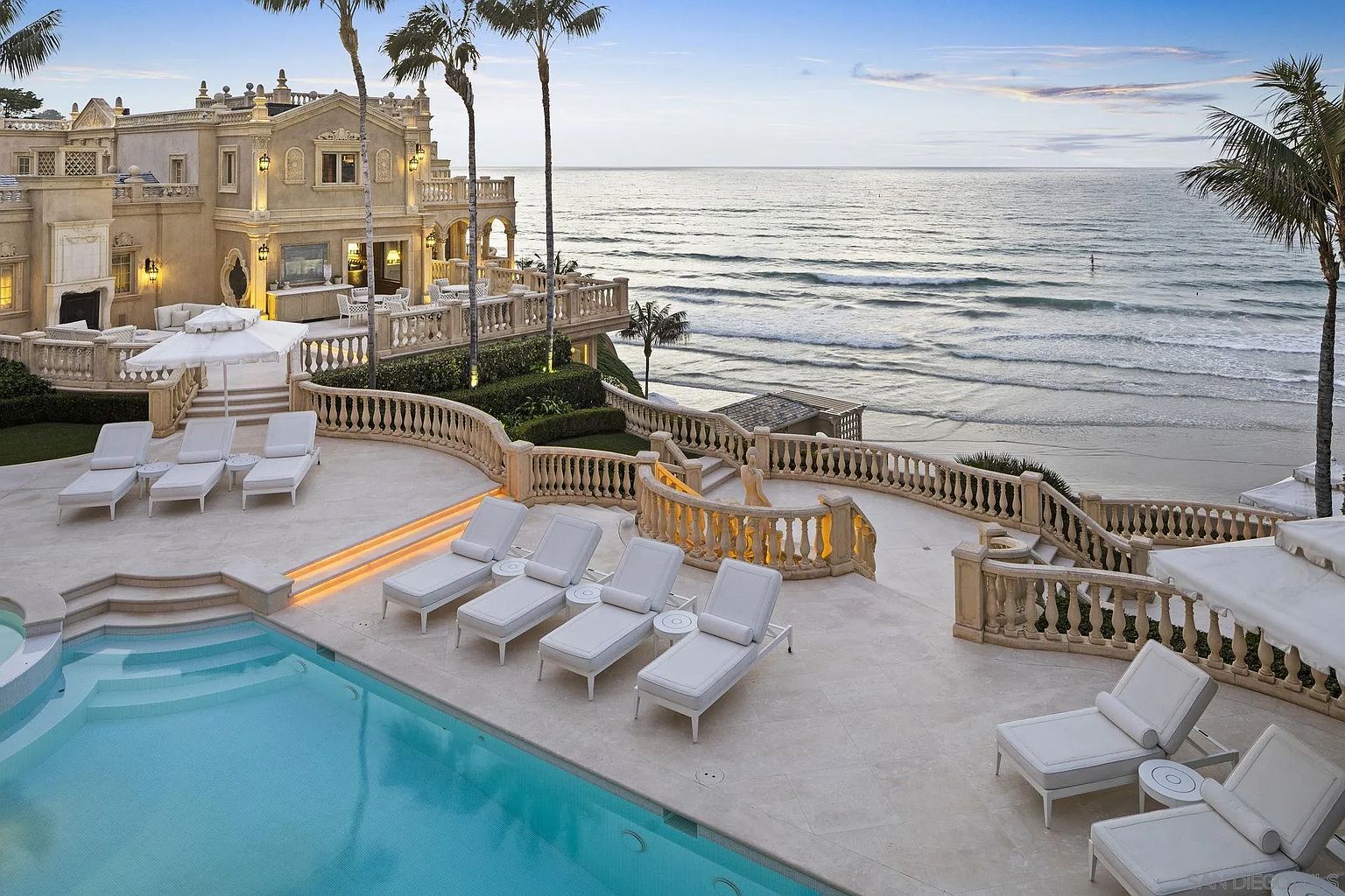 This exterior shot showcases a luxurious pool area with multiple lounge chairs, a pristine pool, and a stunning ocean view. The property features elegant architectural details, including balustrades and manicured landscaping, creating a serene and upscale atmosphere. The image highlights the property's prime location and high-end amenities.