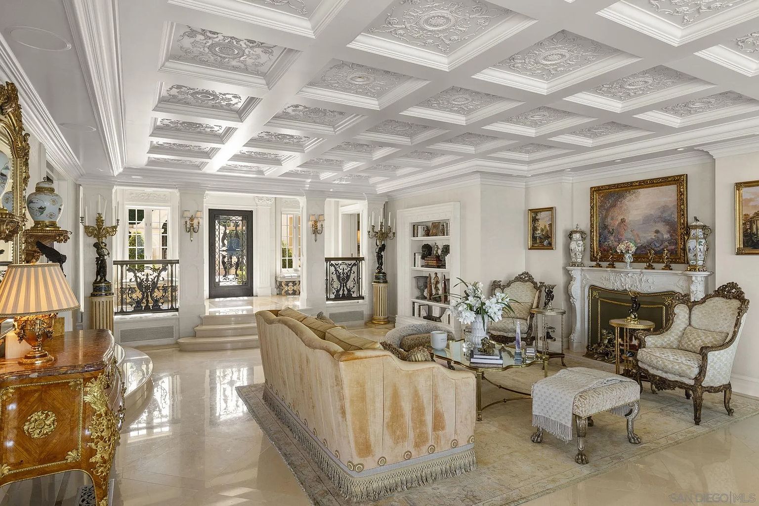 This is an opulent living room featuring a coffered ceiling with intricate detailing, marble flooring, and elegant furnishings. The room is bathed in natural light, highlighting the luxurious finishes and creating a grand, inviting atmosphere. A fireplace with ornate detailing serves as a focal point, complemented by classic artwork and decorative vases.