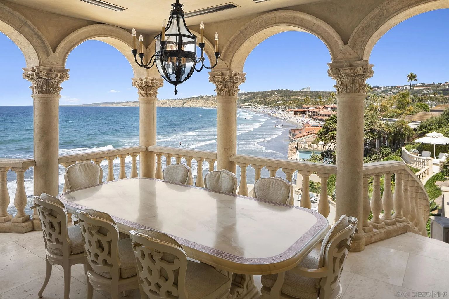 This image showcases a luxurious outdoor dining area on a balcony with stunning ocean views. The space features a large marble dining table surrounded by ornate chairs, all set within an architectural framework of arches and columns. A decorative chandelier hangs above the table, adding to the elegant ambiance.