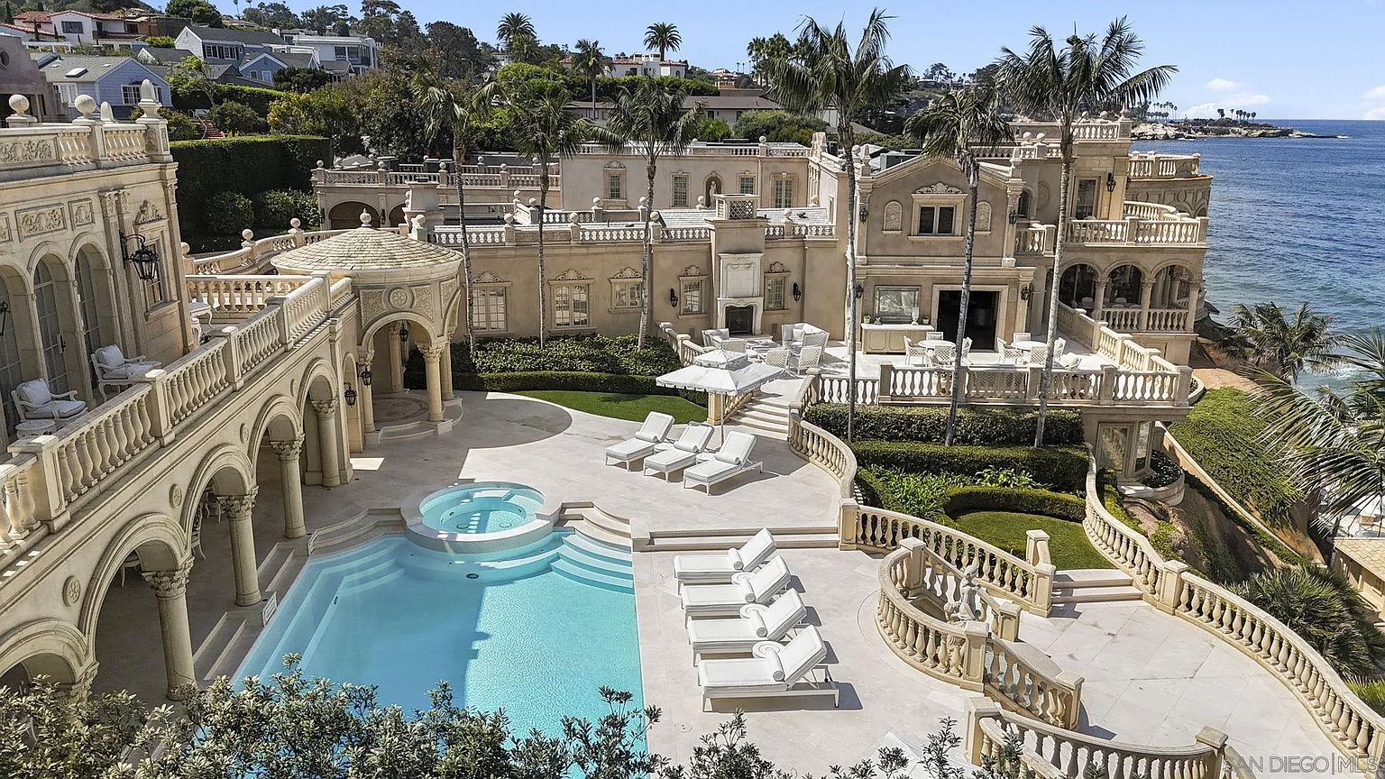 This aerial view showcases a luxurious estate with a stunning pool and spa area. The property features Mediterranean-style architecture with arched walkways, multiple balconies, and meticulously landscaped grounds. The pool is surrounded by lounge chairs, creating an inviting outdoor oasis with ocean views.