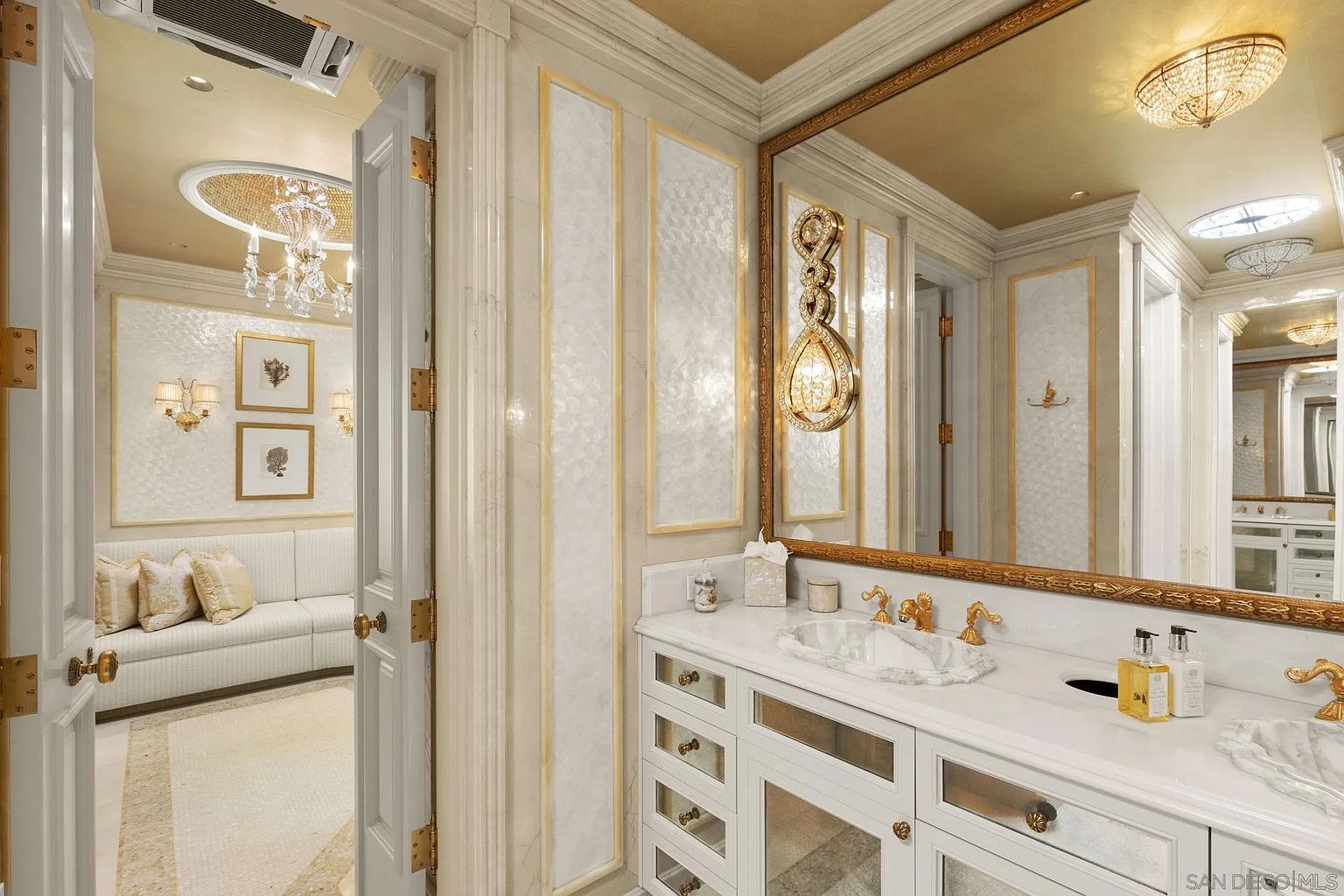 This is a luxurious primary bathroom featuring a double vanity with marble countertops and gold fixtures. The walls are adorned with decorative panels, and a large mirror reflects the opulent lighting and gold-leafed ceiling. The space exudes elegance and sophistication, creating a spa-like atmosphere.