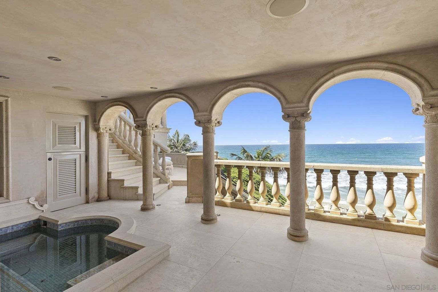 This image showcases a luxurious outdoor balcony featuring elegant architectural details such as stone columns, arched openings, and a decorative balustrade. The balcony overlooks a stunning ocean view with clear blue skies, creating a serene and upscale ambiance. A small pool or spa is visible on the left, adding to the property's appeal.