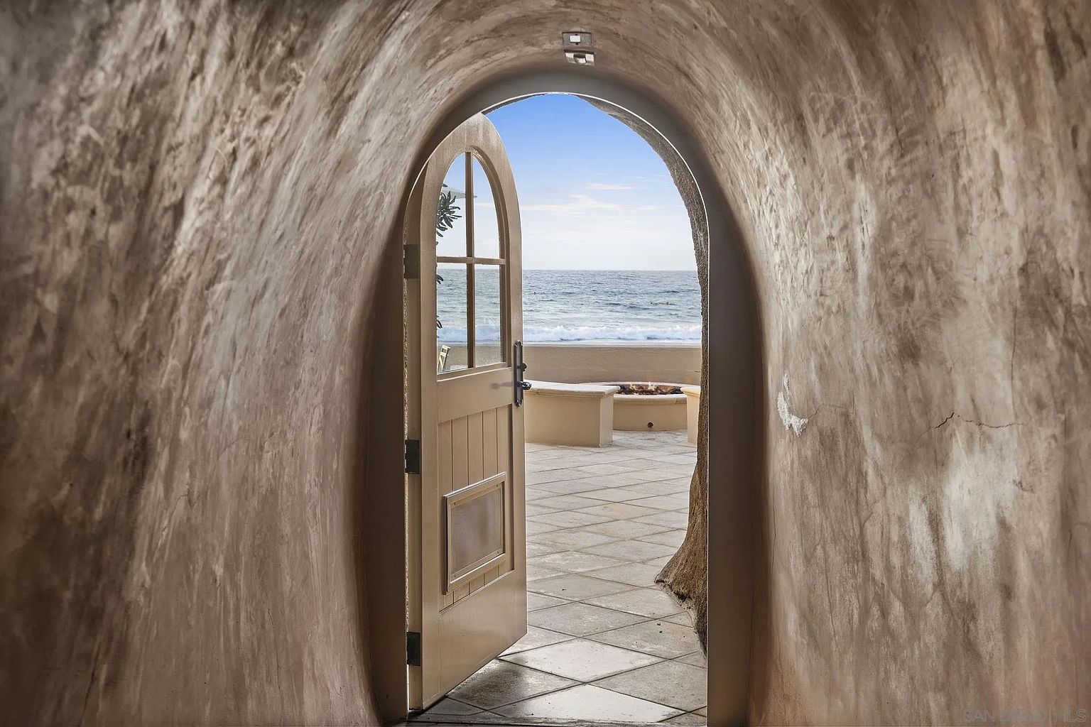 This image showcases a unique patio or balcony space accessible through an arched doorway. The doorway is framed by textured walls, leading to an outdoor area with tiled flooring, a built-in seating area, and a fire pit, all overlooking the ocean. The scene evokes a sense of secluded luxury and relaxation.