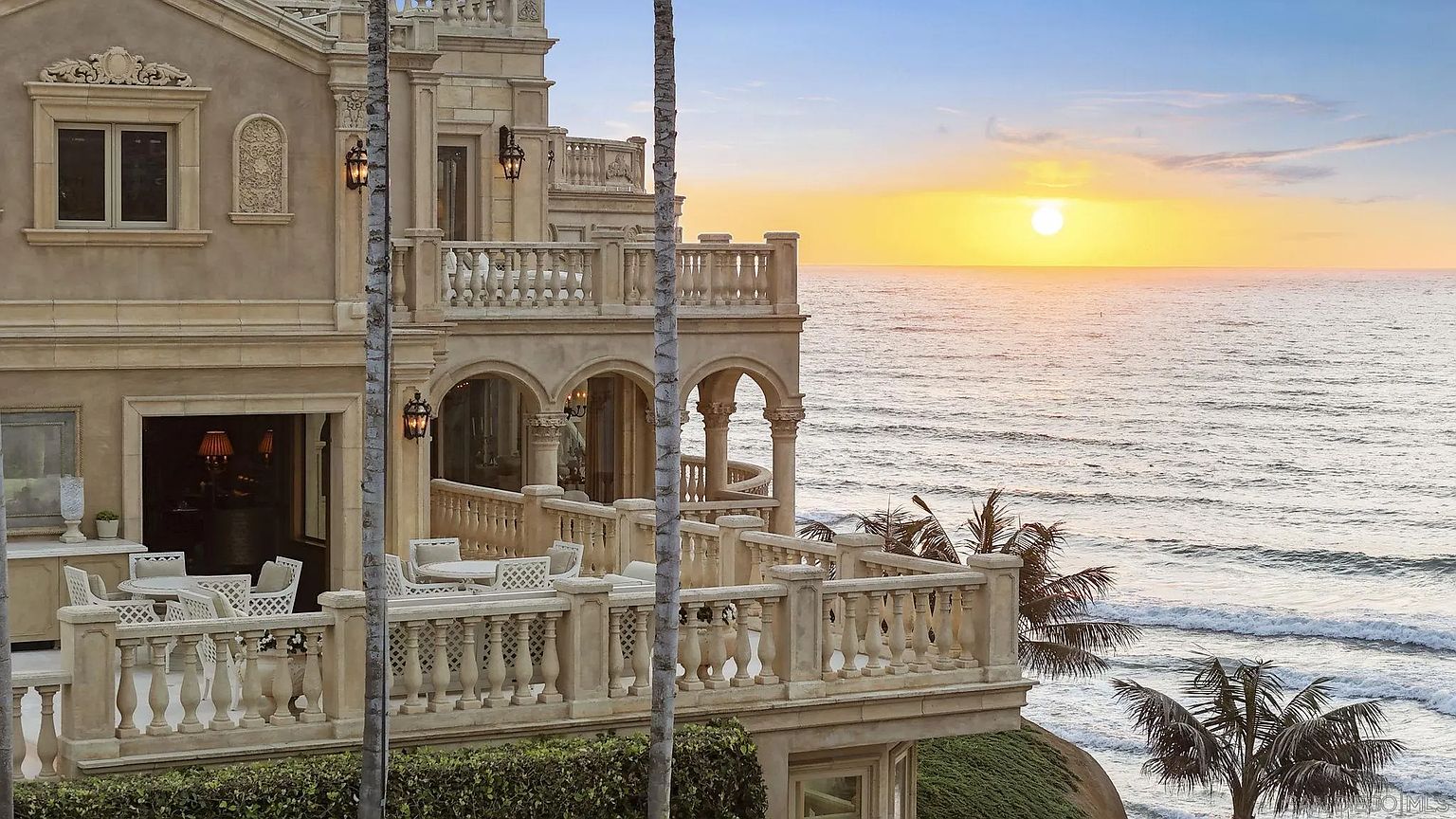 This image showcases a luxurious multi-level patio/balcony area of a grand coastal estate. The architecture features ornate stone balustrades, arched openings, and elegant lighting fixtures. The patio is furnished with outdoor dining sets, offering stunning ocean views at sunset, enhancing the property's appeal for outdoor living and entertaining.