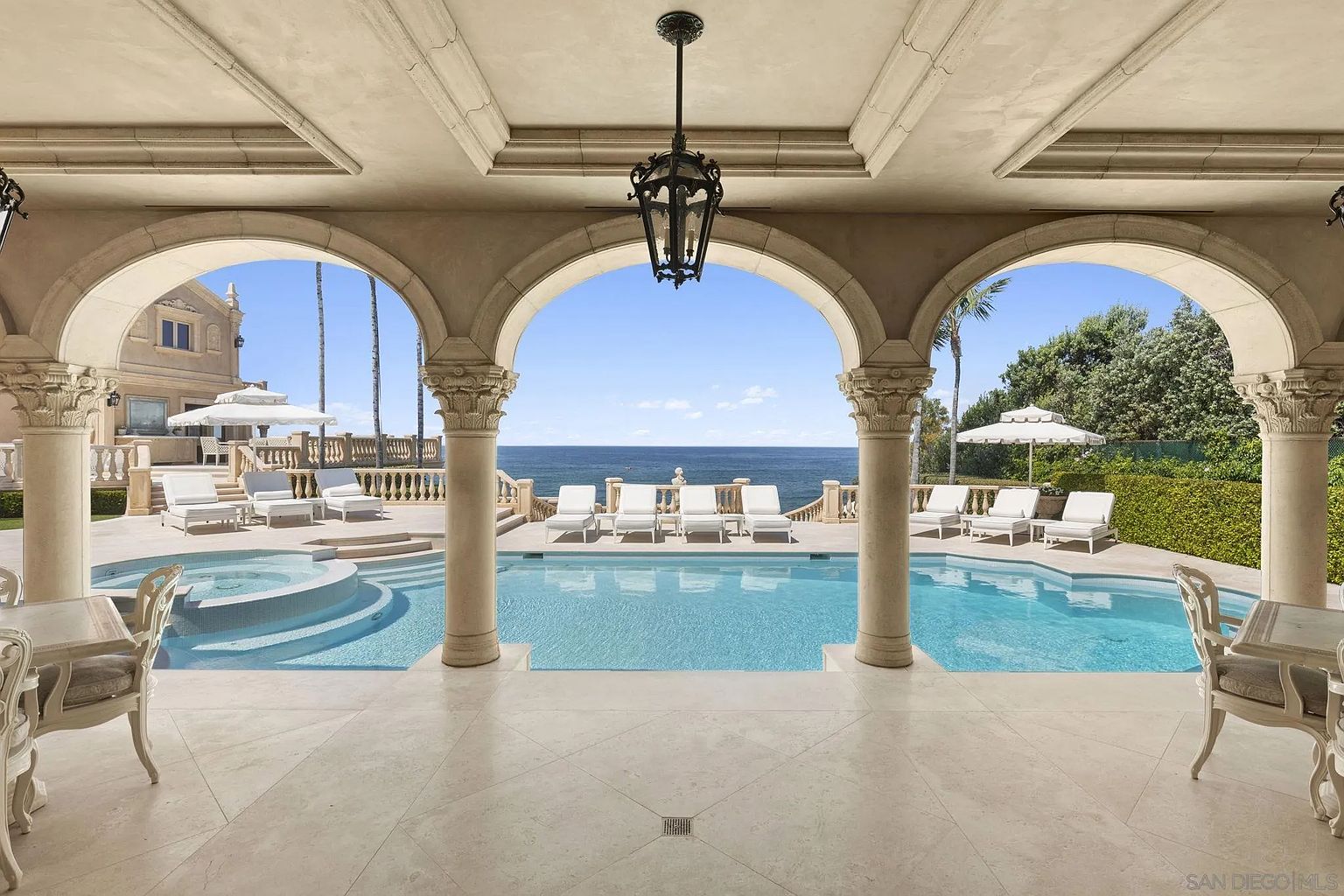 This image showcases a luxurious outdoor pool and spa area, framed by elegant arched columns and a covered patio. The pool is surrounded by lounge chairs, offering a relaxing space with ocean views. The architectural style is classic and sophisticated, creating an inviting and upscale ambiance.