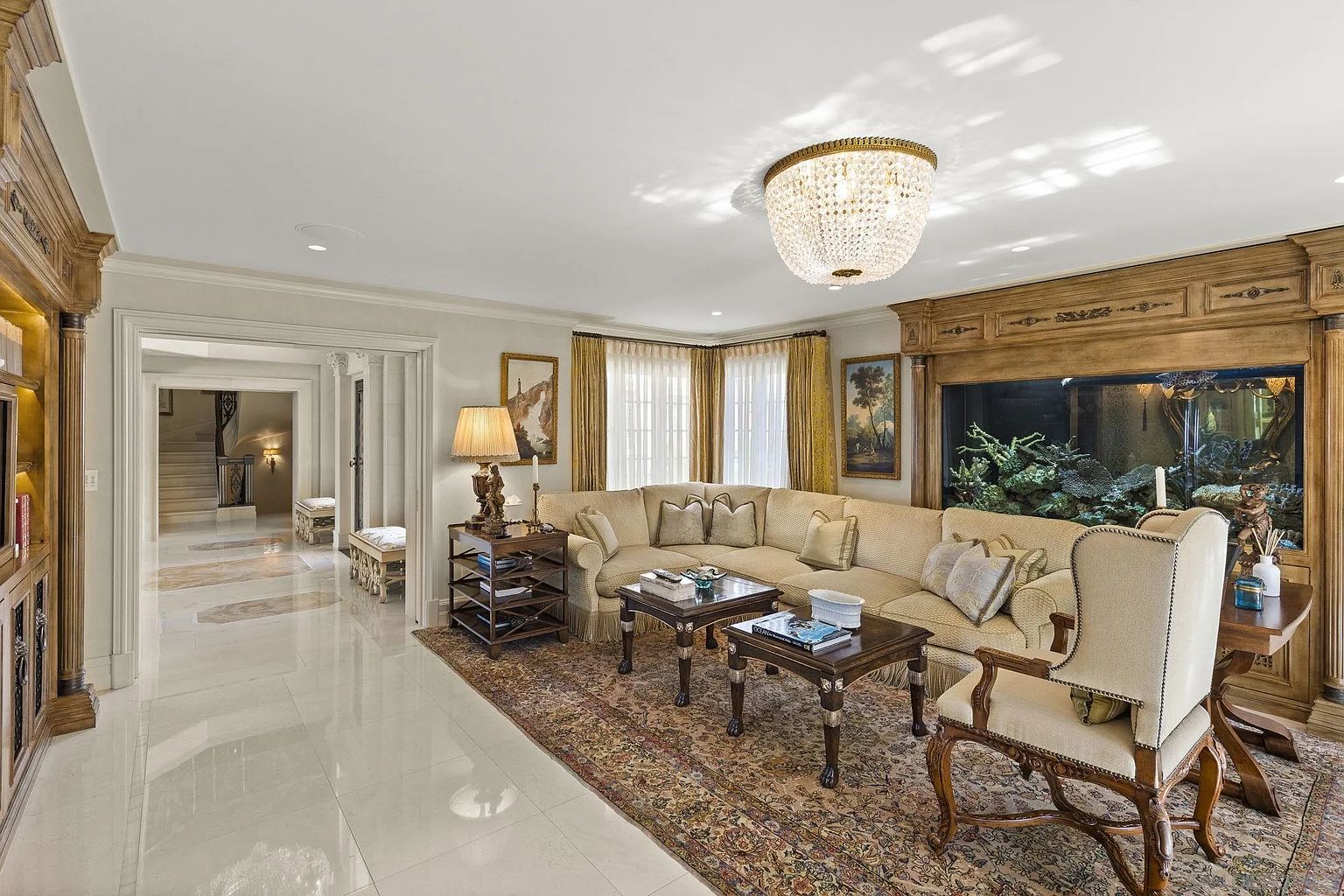 This is an elegant living room featuring a large, comfortable sectional sofa, two dark wood coffee tables, and a decorative area rug. A striking chandelier hangs from the ceiling, and a built-in aquarium adds a unique focal point. The room exudes a luxurious and inviting atmosphere, perfect for relaxation and entertaining.