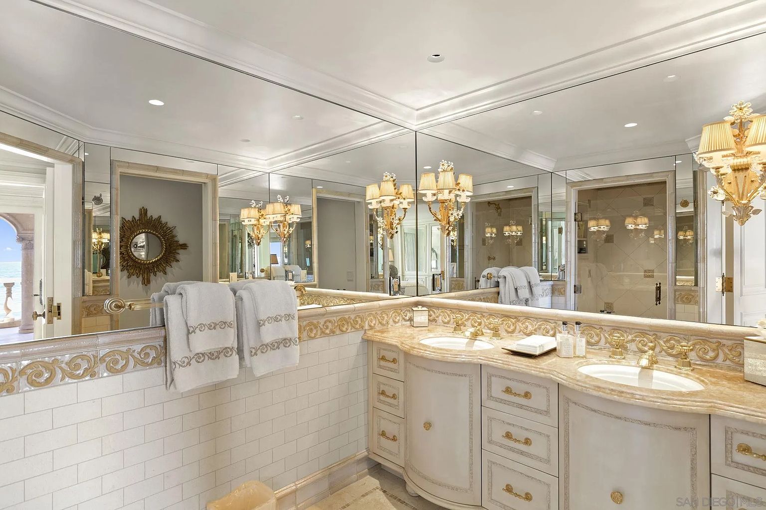 This is a luxurious primary bathroom featuring a double vanity with a marble countertop and ornate gold detailing. The walls are tiled, and a large mirror spans the length of the vanity, reflecting the elegant chandeliers and a glimpse into an adjacent room. The overall impression is one of opulence and sophistication.
